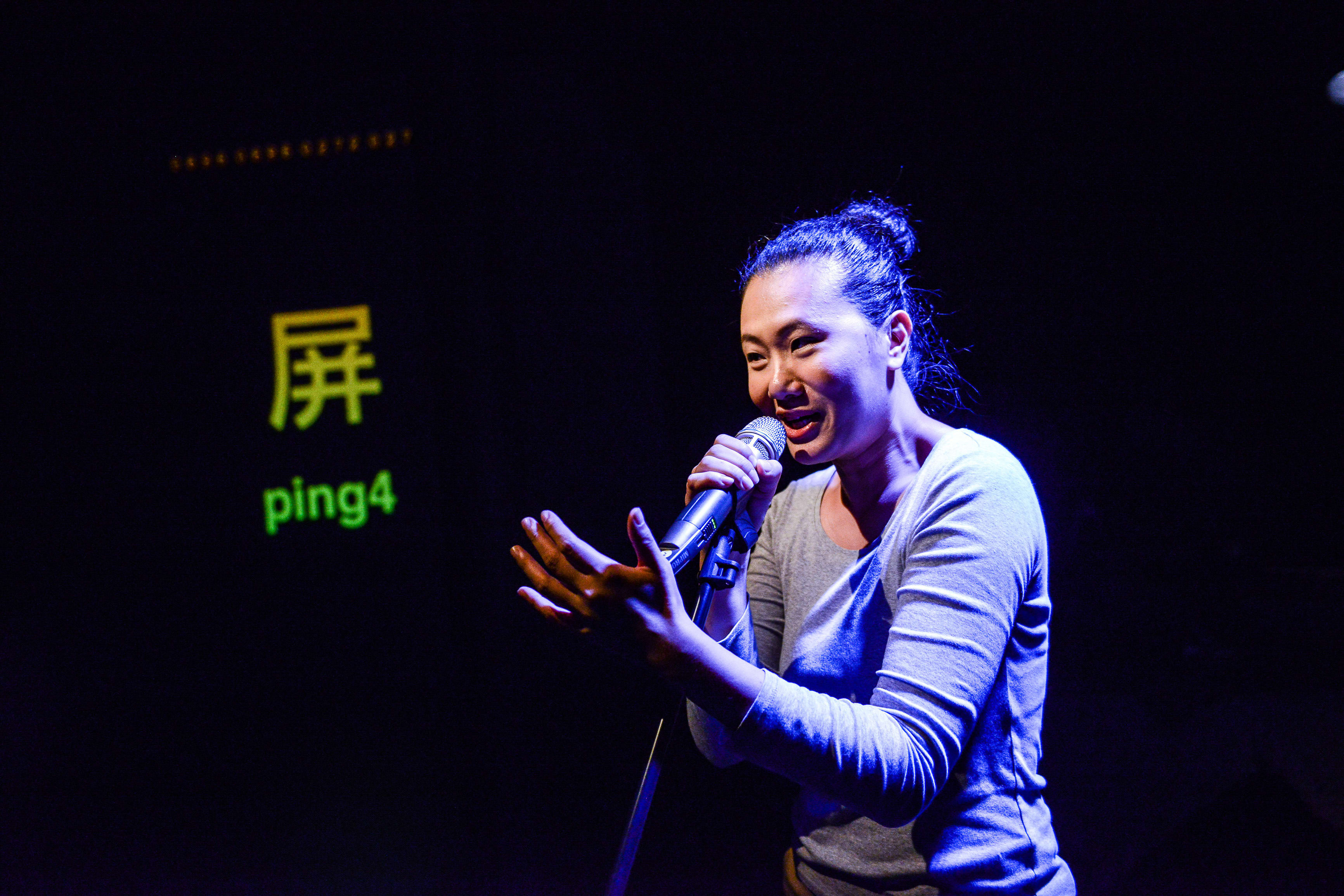 Performer Michelle Li on stage for Rooftop Productions' contemporary theatre piece 'Testimony' ('山下的證詞'). Bathed in dramatic blue spotlight, she holds a microphone and gestures expressively while speaking to the audience. Behind her, a dark screen displays the Chinese character '屏' and the Jyutping text 'ping4'. Michelle Li delivers a poignant line from the production: "So to learn Cantonese, you have to learn Jyutping."
