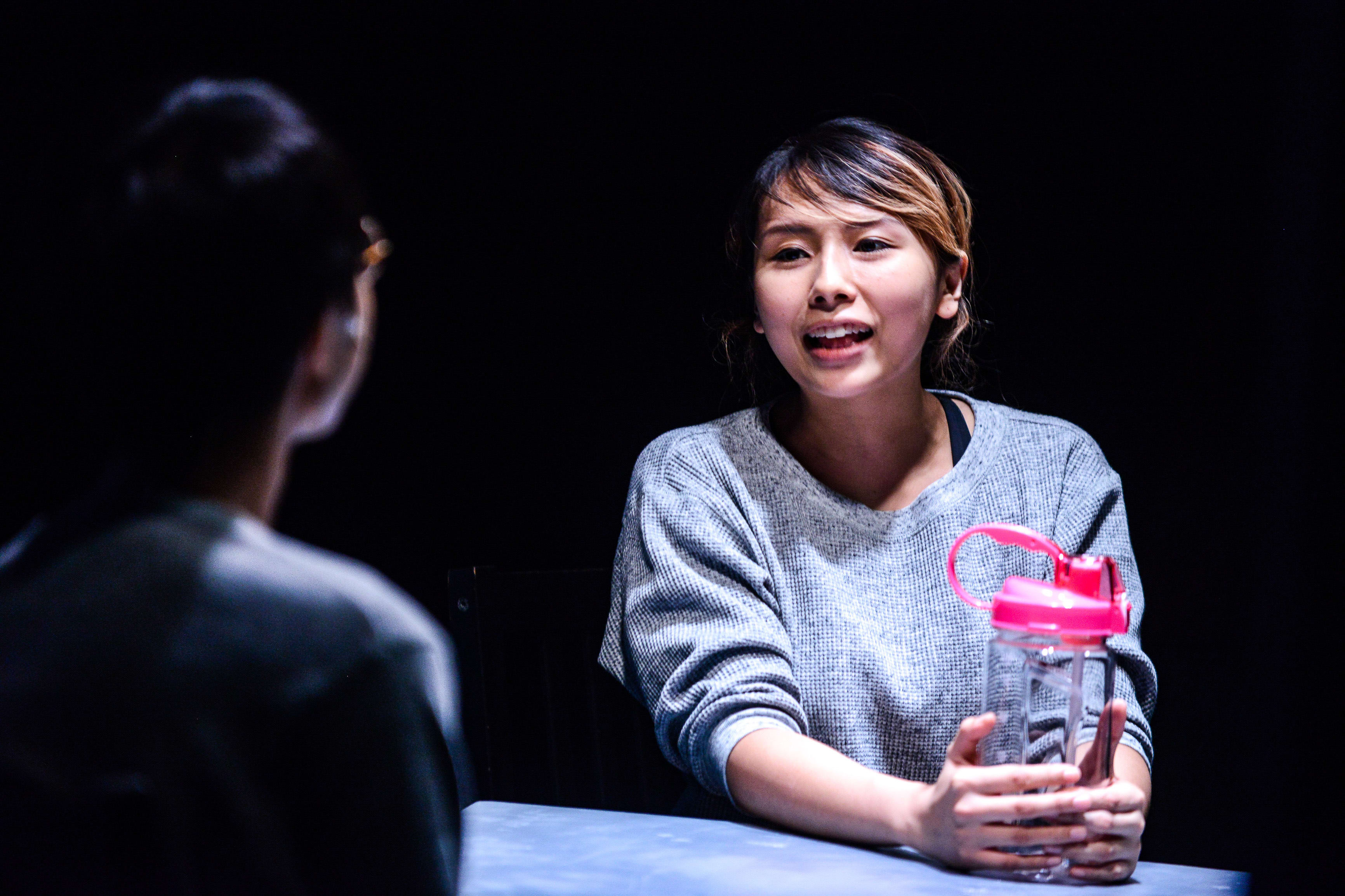 天台製作的當代香港劇場作品《山下的證詞》（Testimony）中，演員李里莎（Arisa Yasaratne）在漆黑的舞台背景下，被聚光燈照亮，手持水瓶神情專注地進行演出。「但是我身邊大部分的同學，也是在讀本地小一的中文。」