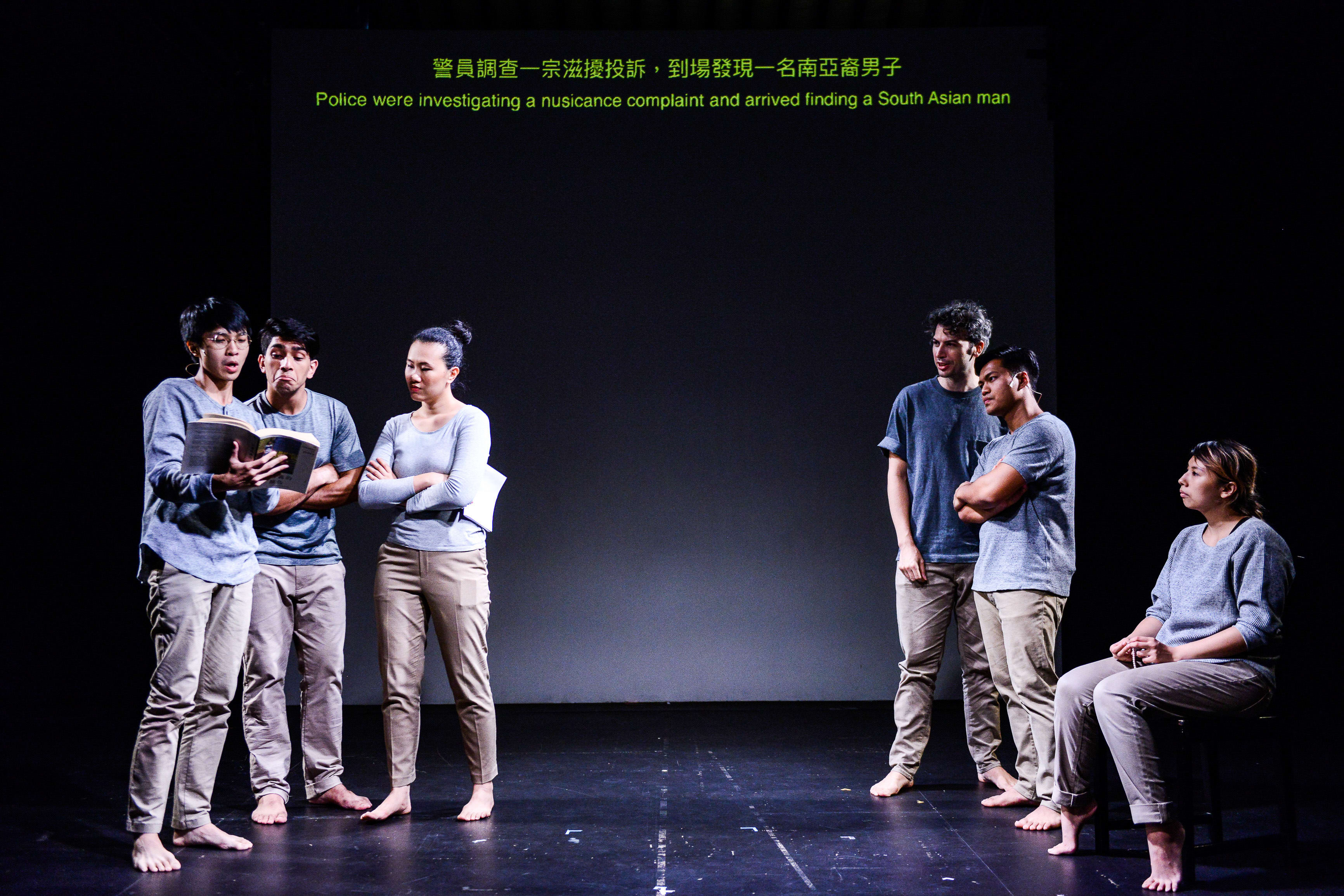 In a dramatic scene from Rooftop Productions' contemporary theatre piece 'Testimony' (《山下的證詞》), five actors are staged on a stark, minimalist black stage. The lighting highlights a central dialogue, with a screen above displaying text about a police nuisance complaint involving a South Asian man. The performance explores complex social themes in Hong Kong through powerful physical theatre. One performer asks, "Tusar, do you know what the Limbu case is?"