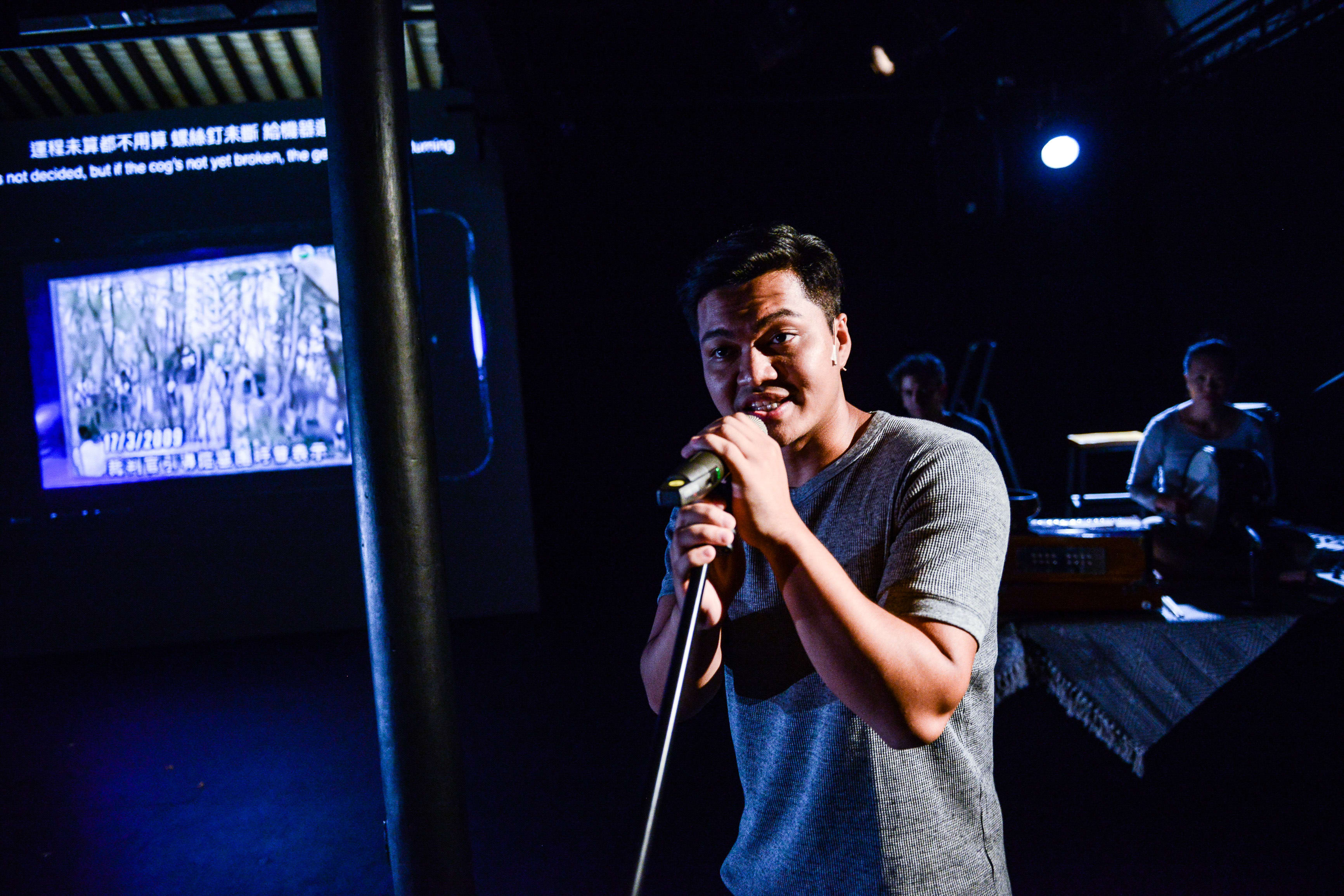 Basnet Tusar delivers a poignant performance in Rooftop Productions' contemporary Hong Kong theatre piece, 'Testimony' (山下的證詞). He stands under dramatic, moody stage lighting, holding a microphone with an intense, focused expression. In the background, a projection screen displays a historical, grainy image of a forest scene dated 17/3/2009. 'Testimony'