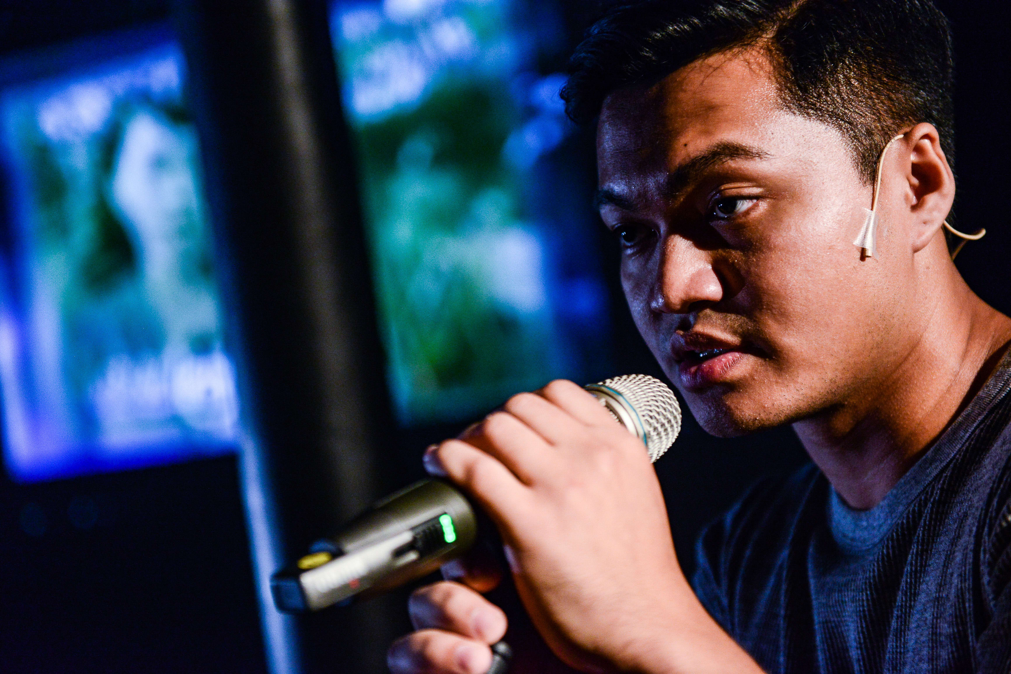 演員 Basnet Tusar 於天台製作（Rooftop Productions）當代香港劇場作品《山下的證詞》演出期間，在充滿戲劇感的藍色燈光背景下，神情凝重地手持麥克風發言。這張特寫鏡頭捕捉了角色訴說劇中台詞的動人瞬間：「造就你嗅不到的景氣」。