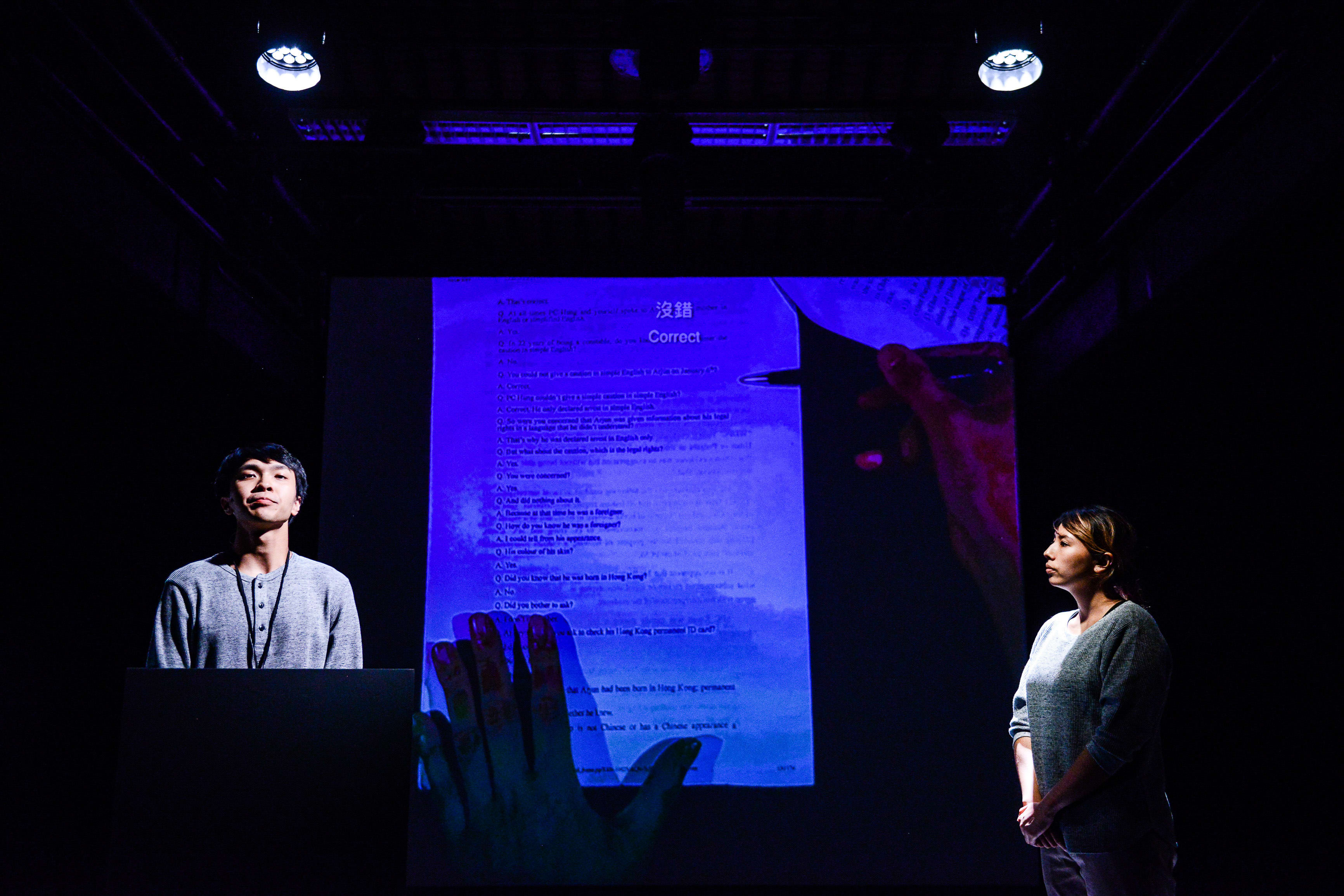 On a dark stage lit by two overhead spotlights, actors Ng King Lung and Arisa Yasaratne stand on opposite sides of a large, glowing projection of an exam paper. Ng King Lung appears on the left, while Arisa Yasaratne stands on the right, both framed by the blue-tinted, semi-transparent image of a hand marking a test as 'Correct.' This scene from the Rooftop Productions contemporary theatre performance 'Testimony' highlights themes of memory and scrutiny. 'Testimony'