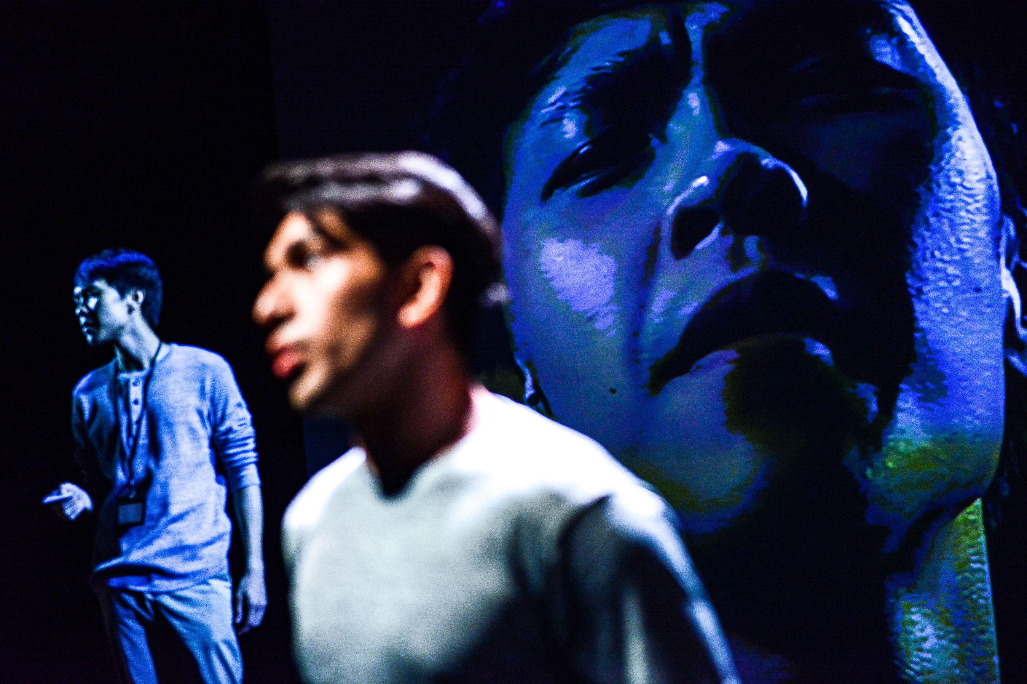 In a dramatic scene from Rooftop Productions' contemporary theatre piece 'Testimony', actor Mohammad Kashif Ali stands in the foreground, his expression intense and pained. Behind him, another performer is softly illuminated in the shadows, while a colossal, high-contrast blue-toned projection of a face looms over the stage, adding a haunting, immersive layer to this Hong Kong stage performance. Mohammad Kashif Ali speaks the line: "So the first time I got checked it was downstairs at my home"
