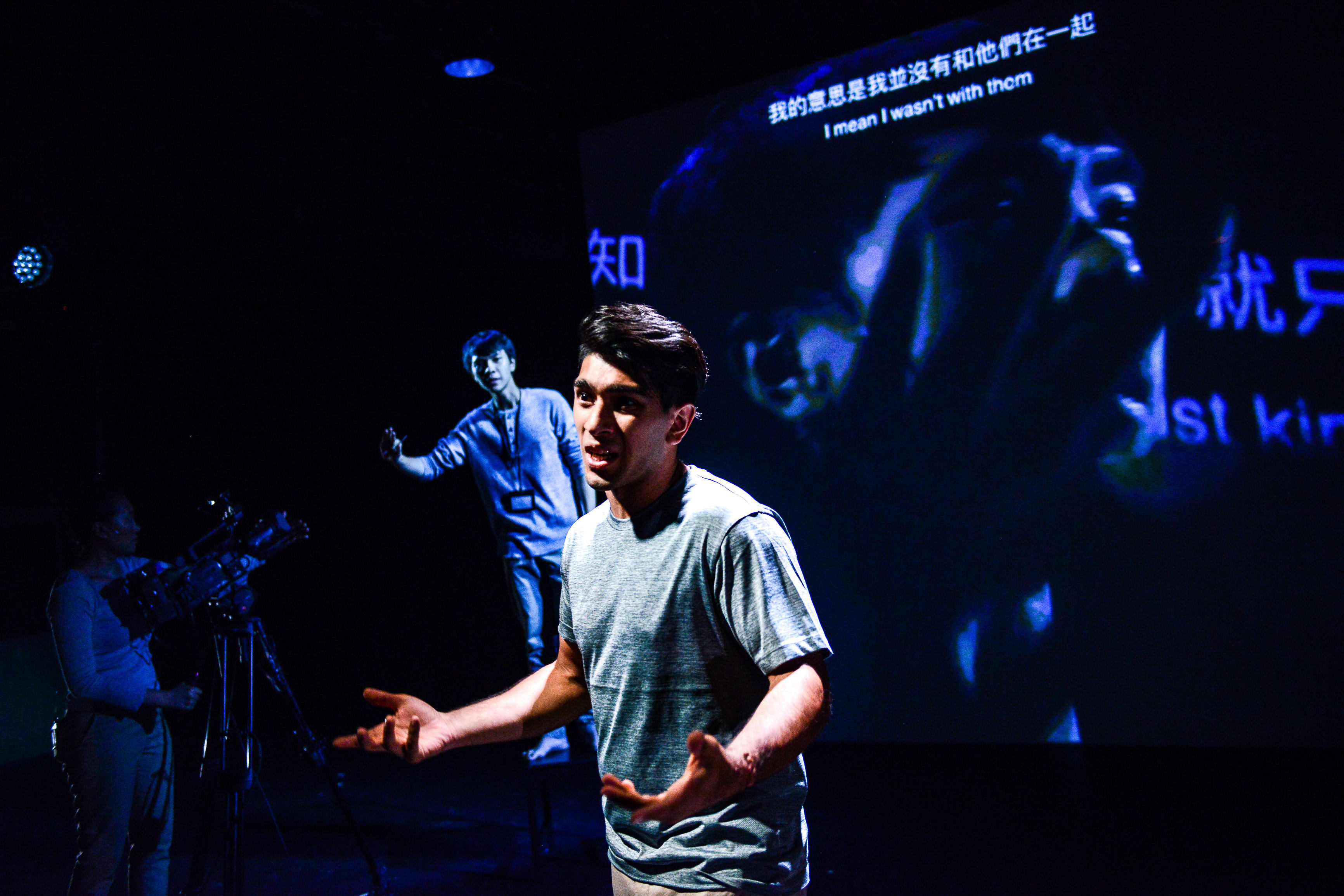 In a dramatic scene from Rooftop Productions' contemporary theatre piece 'Testimony' (《山下的證詞》), actor Mohammad Kashif Ali (巫加沙) stands center stage under sharp, low-key lighting, gesturing expressively as he delivers a monologue. Behind him, another performer stands on a raised platform, while a large projection displays the translated text: 'I mean I wasn't with them'. A camera operator is visible on the left, highlighting the multimedia nature of this Hong Kong stage production. As the performer speaks, he declares: 'Testimony'