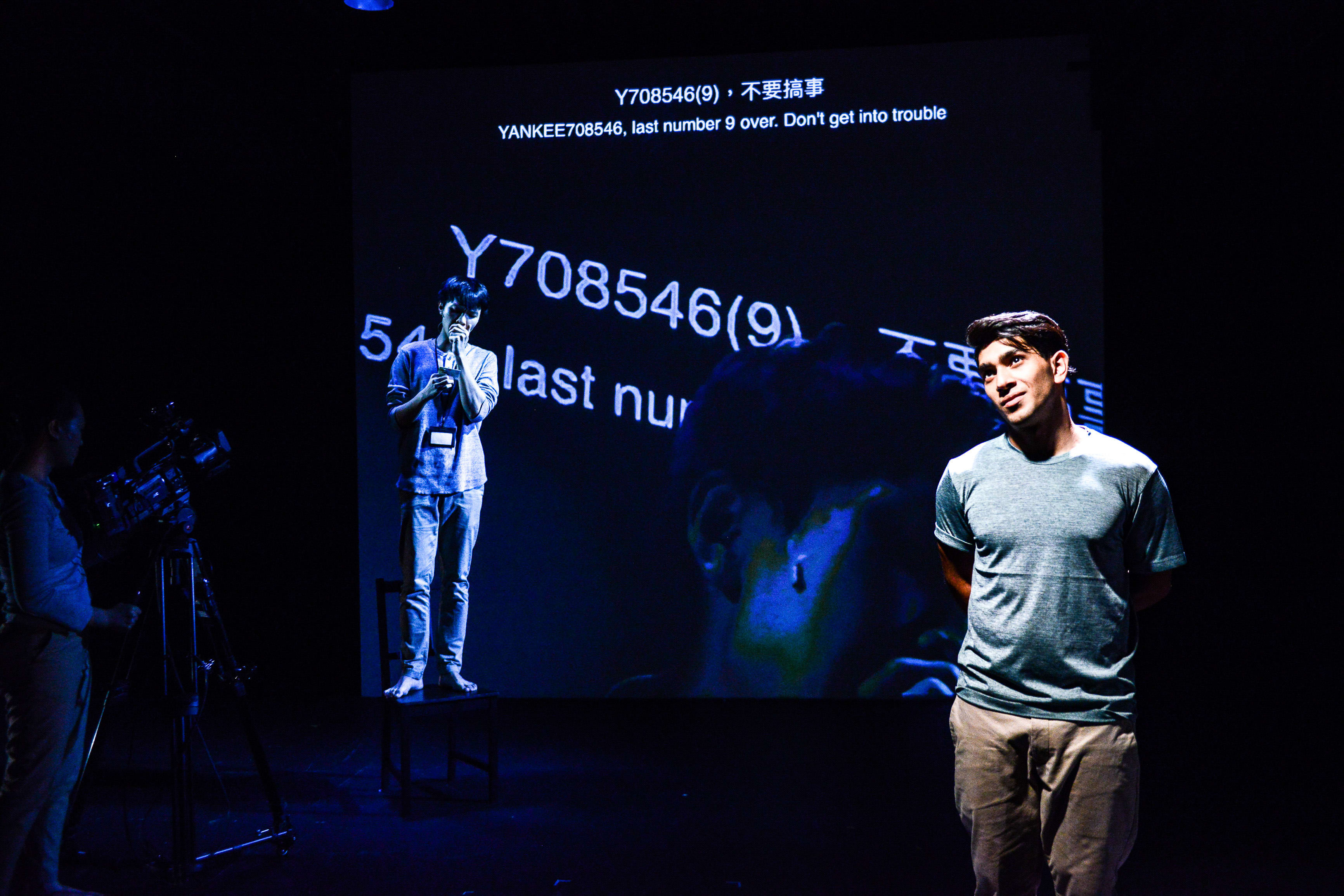 天台製作（Rooftop Productions）現代劇場作品《山下的證詞》（Testimony）的舞台演出劇照。在昏暗的燈光下，演員吳景隆站在椅子上，神情專注地望向身後的大型投影幕，螢幕上顯示著「YANKEE708546(9)，不要搞事」的字樣。巫加沙（Mohammad Kashif Ali）則站在前景，雙手背在身後，若有所思地看向舞台側方。台上演員說道：「總而言之，這是我跟警察唯一的有趣經歷。」