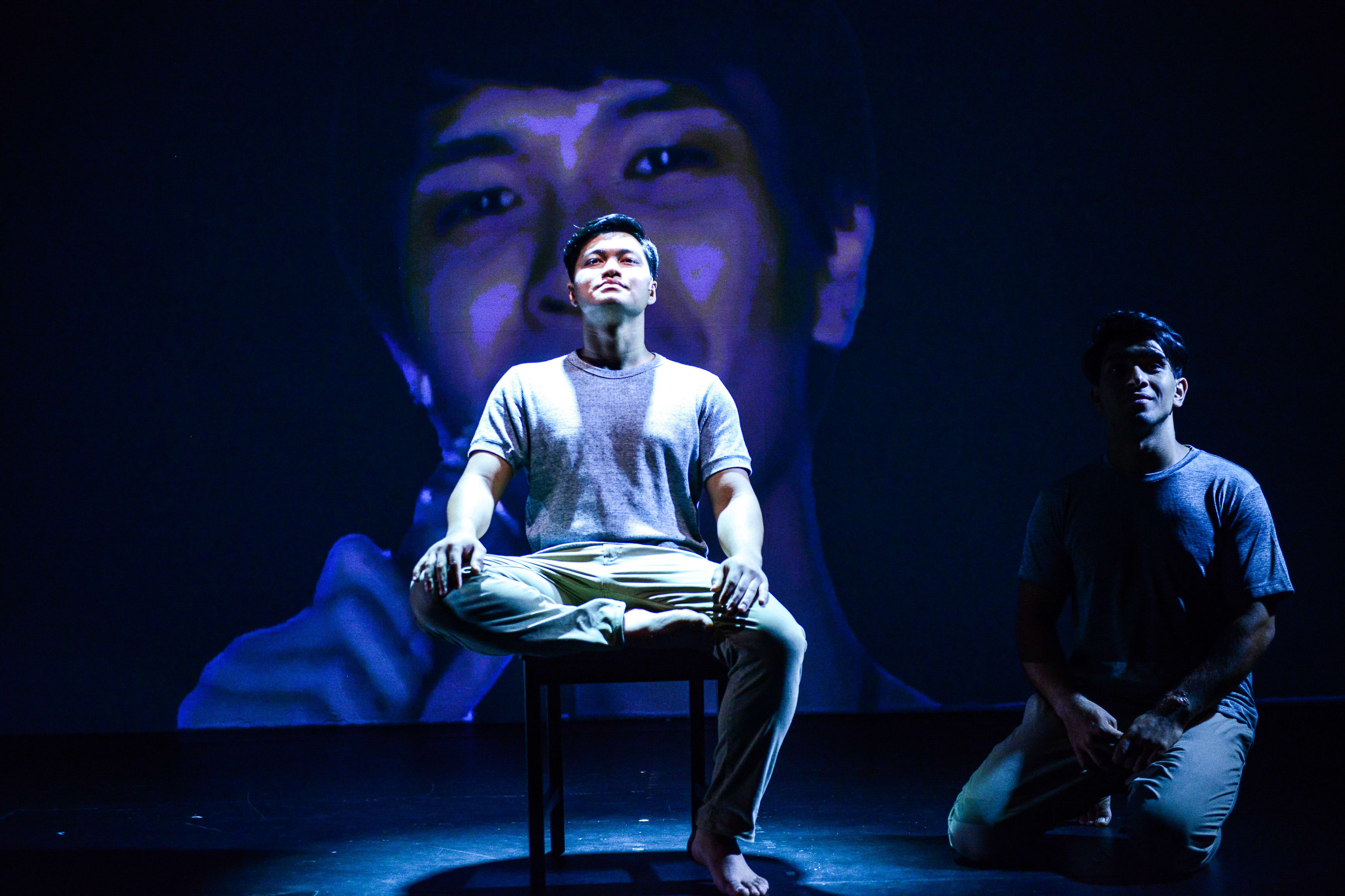 在天台製作（Rooftop Productions）的當代劇場作品《山下的證詞》（Testimony）中，一名演員在舞台中央的椅子上盤腿而坐，沐浴在強烈的藍色聚光燈下。他身後巨大的投影映出他模糊的臉龐，呼應著沉思的神情。舞台右側，另一位演員跪在陰影中，同樣被冷調的藍光籠罩，為這場香港劇場演出營造出充滿戲劇張力的氛圍。《山下的證詞》