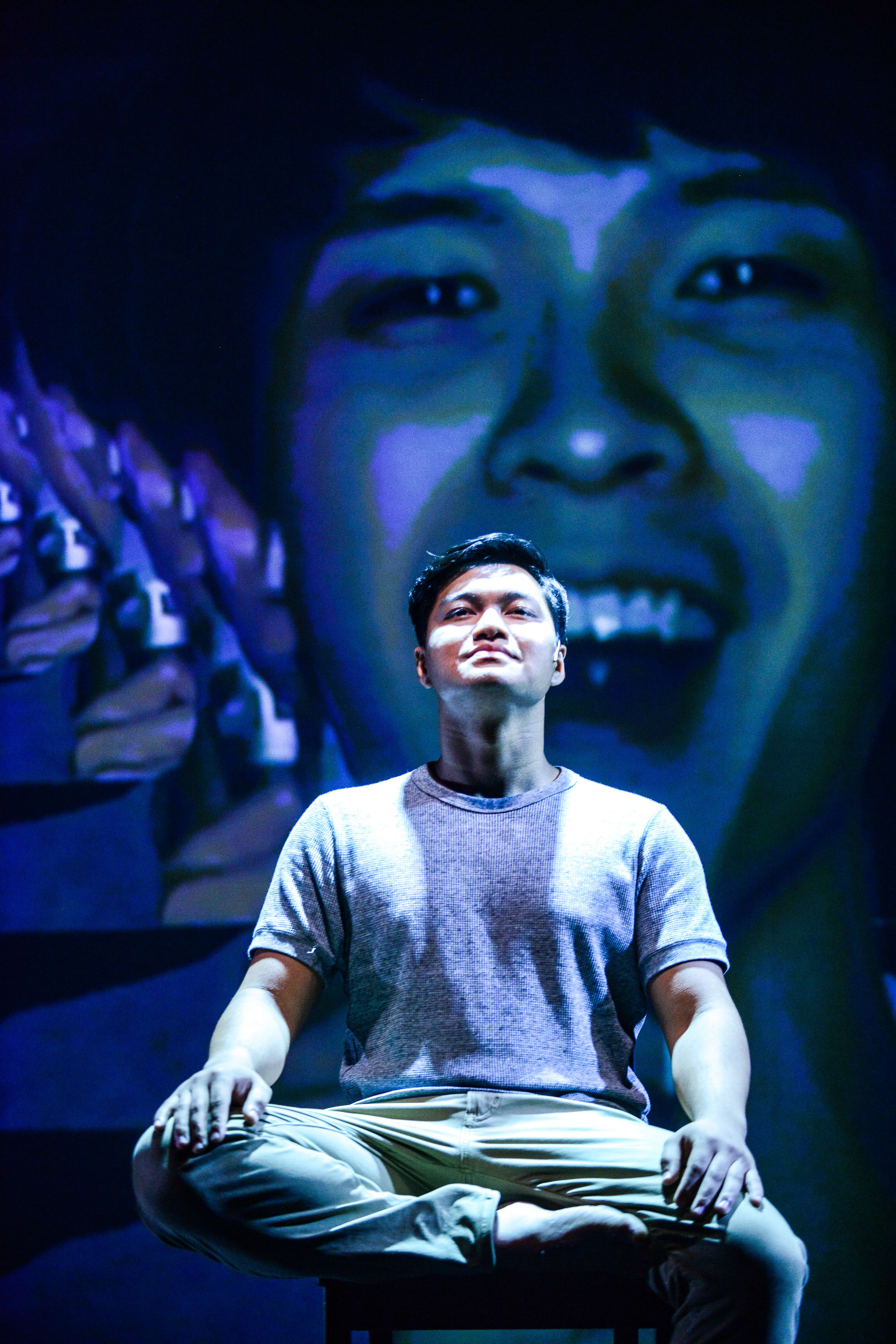 Basnet Tusar sits in a cross-legged meditative pose on a minimalist stool, bathed in a sharp, cool-toned spotlight. Behind him, a massive, out-of-focus projection of his own face dominates the stage, creating a surreal and introspective atmosphere characteristic of Rooftop Productions' contemporary Hong Kong theatre. As he gazes upward with a pensive expression, he delivers the line: "And revolt? Why do men revolt?"