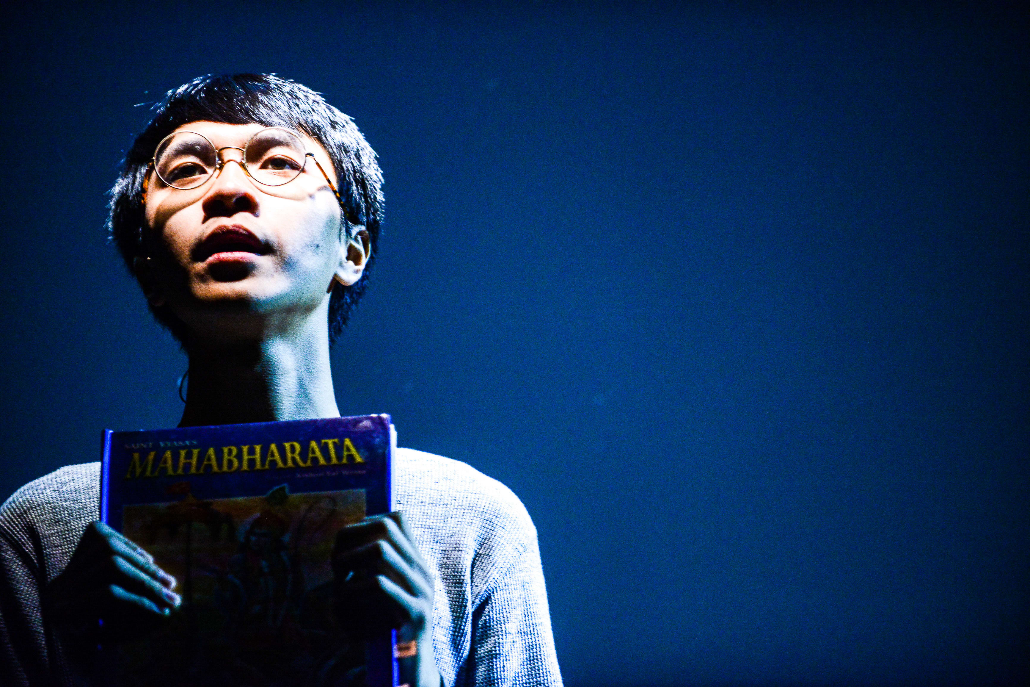 In a moody, blue-lit scene from Rooftop Productions' contemporary Hong Kong play 'Testimony' (《山下的證詞》), actor Ng King Lung stands on stage holding a copy of the 'Mahabharata'. His gaze is directed slightly upward, capturing an expression of profound contemplation and introspection. As he portrays the weight of historical memory on stage, he declares: 'The future does not exist, but I can give you eyes to see the battle'