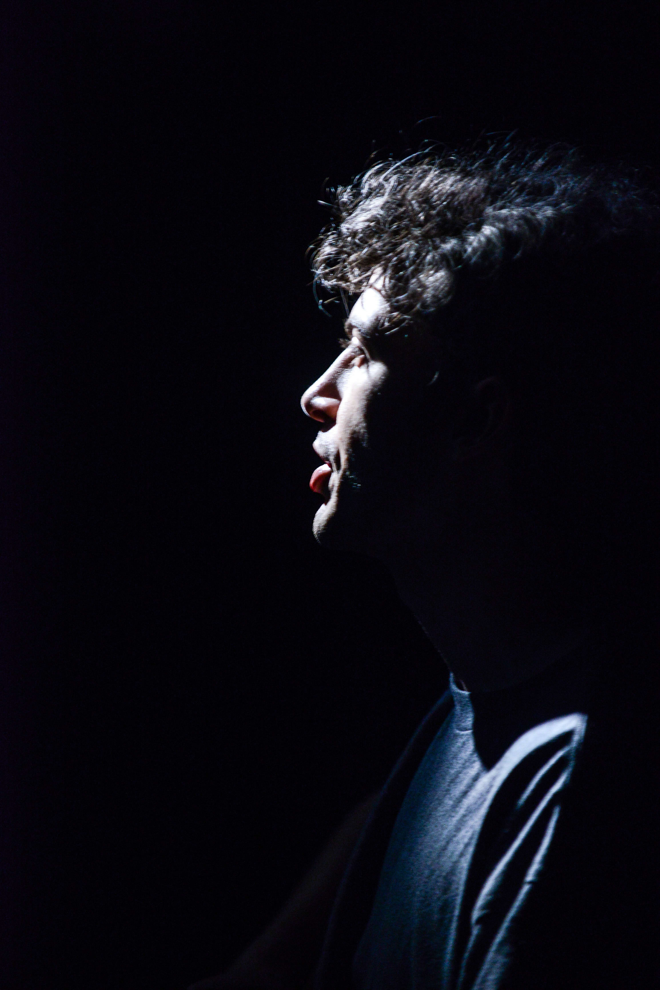 A profile shot of actor Ivor Houlker, illuminated by stark, high-contrast spotlighting against a pitch-black background, capturing an intense and evocative moment from the contemporary Hong Kong theatre production 'Testimony' (《山下的證詞》). The dramatic lighting emphasizes his expression as he performs. 'Testimony'