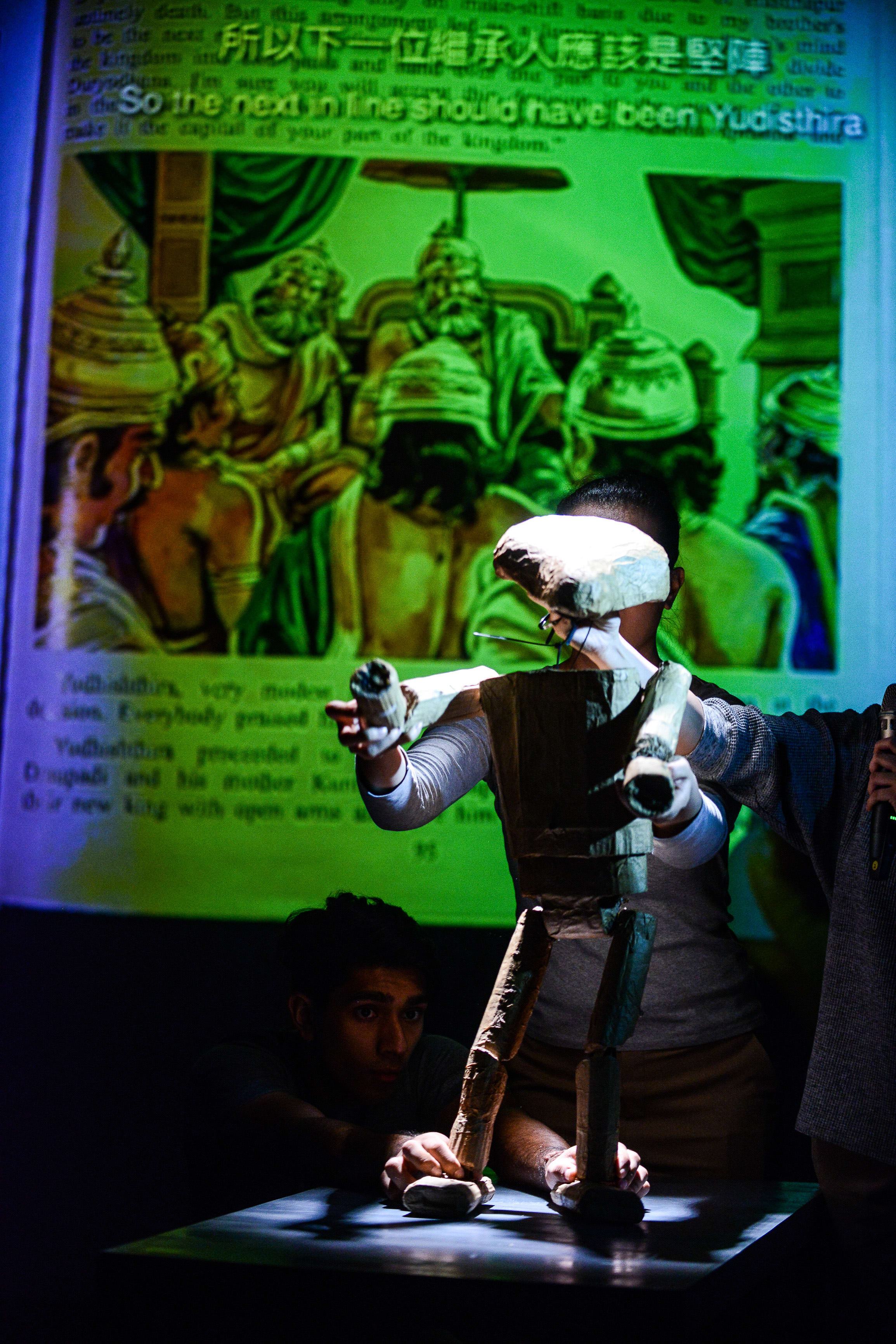 In a scene from Rooftop Productions' contemporary Hong Kong theatre work 'Testimony' (《山下的證詞》), actor Mohammad Kashif Ali operates a wooden puppet on a dimly lit stage. The performance is set against a large-scale projected illustration depicting a scene from the Mahabharata, with text reading, 'So the next in line should have been Yudisthira'. The atmospheric lighting highlights the intimate interaction between the performer and the puppet, capturing the essence of this experimental stage performance.