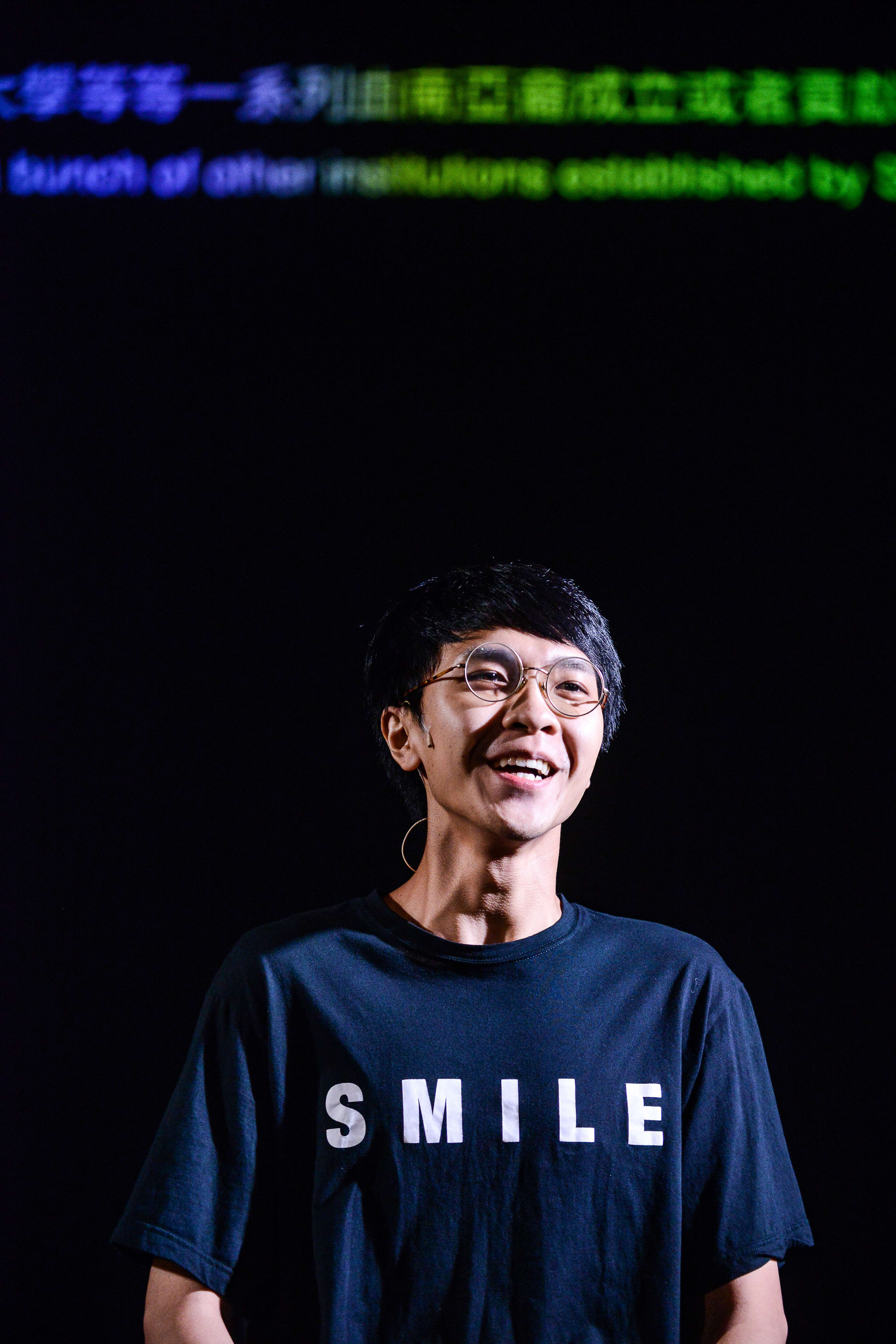 演員吳景隆站在簡約且幽暗的舞台上，聚光燈映照出他充滿感染力的笑容。他身穿一件印有「SMILE」字樣的黑色T恤，展現了香港當代劇場的極簡美學。背景投影出與劇目相關的文字，這張照片捕捉了天台製作（Rooftop Productions）劇目《山下的證詞》中的動人瞬間。《山下的證詞》
