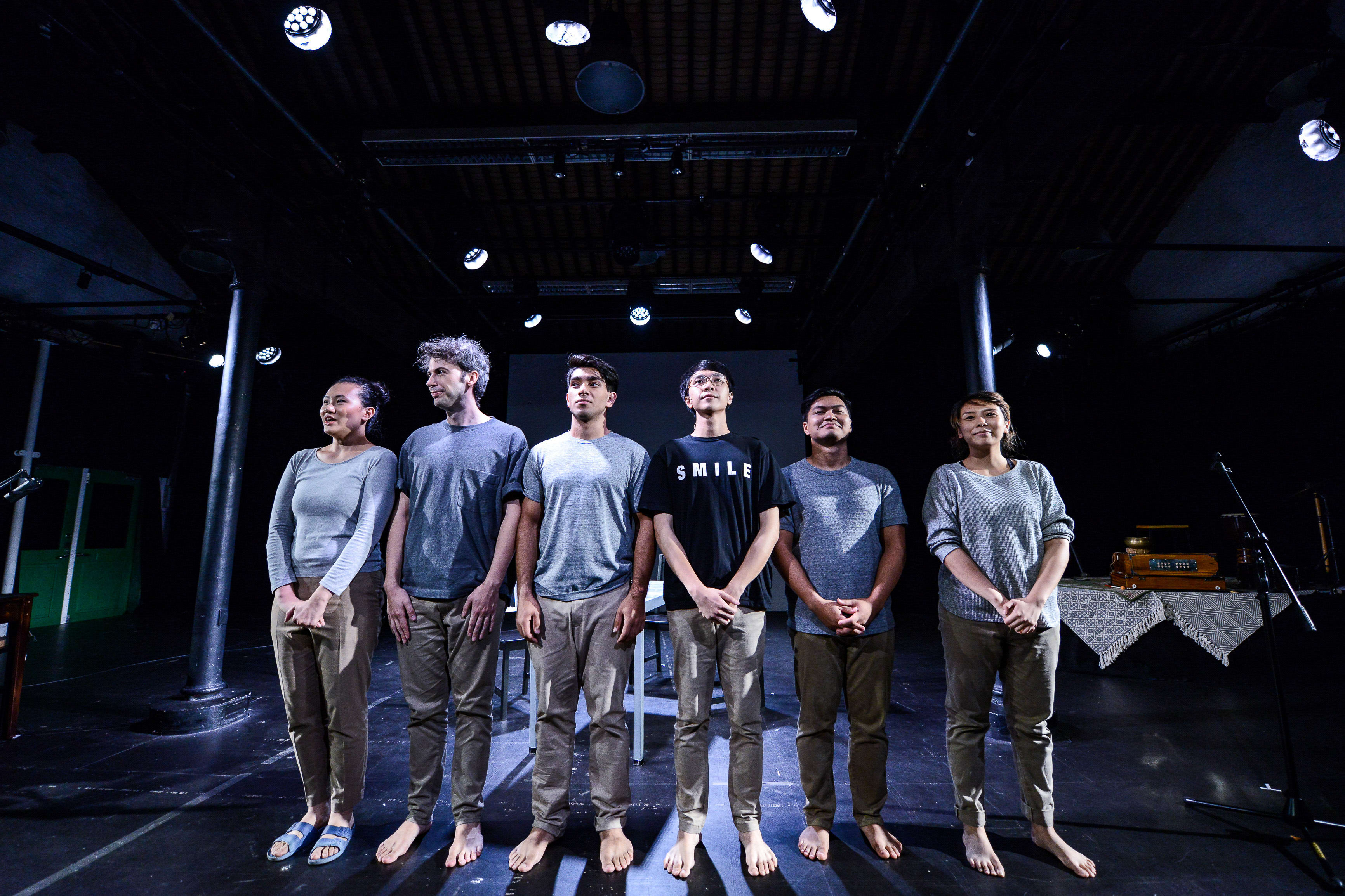 六位演員於天台製作（Rooftop Productions）的當代劇場作品《山下的證詞》舞台上並排站立。演員們穿著大地色系的休閒服裝，在昏暗的劇場燈光下齊齊仰望，神情莊嚴且深沉，呈現出香港當代實驗劇場獨有的情感張力與舞台美學。《山下的證詞》