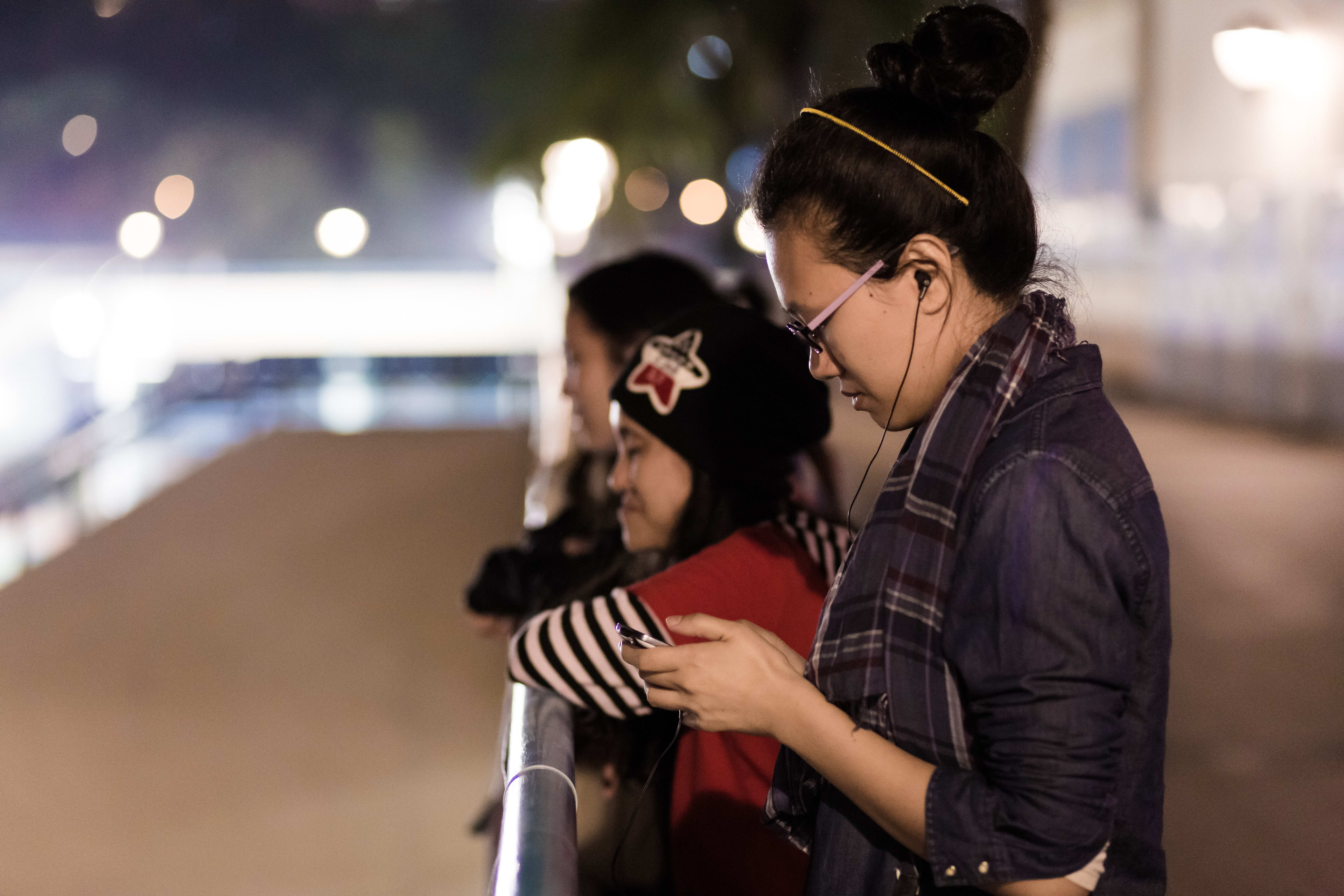 In a candid, low-light shot from Rooftop Productions’ contemporary Hong Kong theatre piece 'The Beautiful Ones' (《行為淪喪》), three women stand along a railing at night. In the foreground, actor Michelle Li (李婉晶) is captured in profile, wearing glasses and earbuds, intently looking down at her mobile phone. Behind her, another performer wearing a beanie with a star emblem gazes toward the cityscape, capturing the reflective, urban intimacy central to this performance. 'The Beautiful Ones'