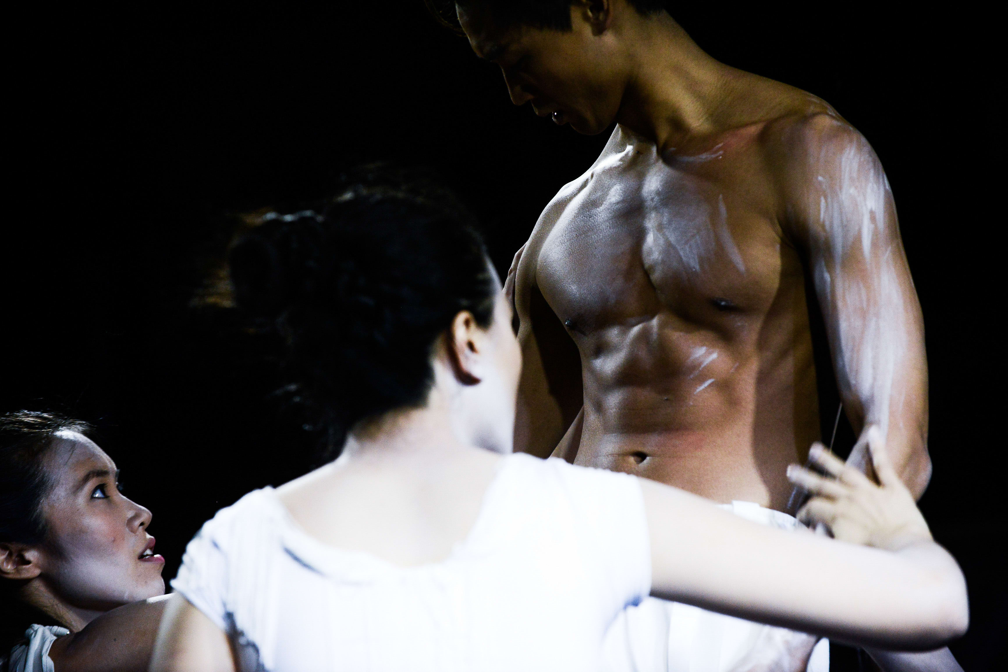 在這張極具張力的劇照中，天台製作（Rooftop Productions）的當代劇場作品《行為淪喪》（The Beautiful Ones）展現了充滿情感張力的一幕。在全黑的背景與強烈的光影對比下，梁皓貽（Isabella Leung）正以充滿情感的眼神凝視著身旁的上身赤裸的男演員；男演員的肌膚上抹有白色的油彩，呈現出獨特的視覺效果。這場演出展現了香港當代劇場對身體表演與舞台美學的深度探索。《行為淪喪》