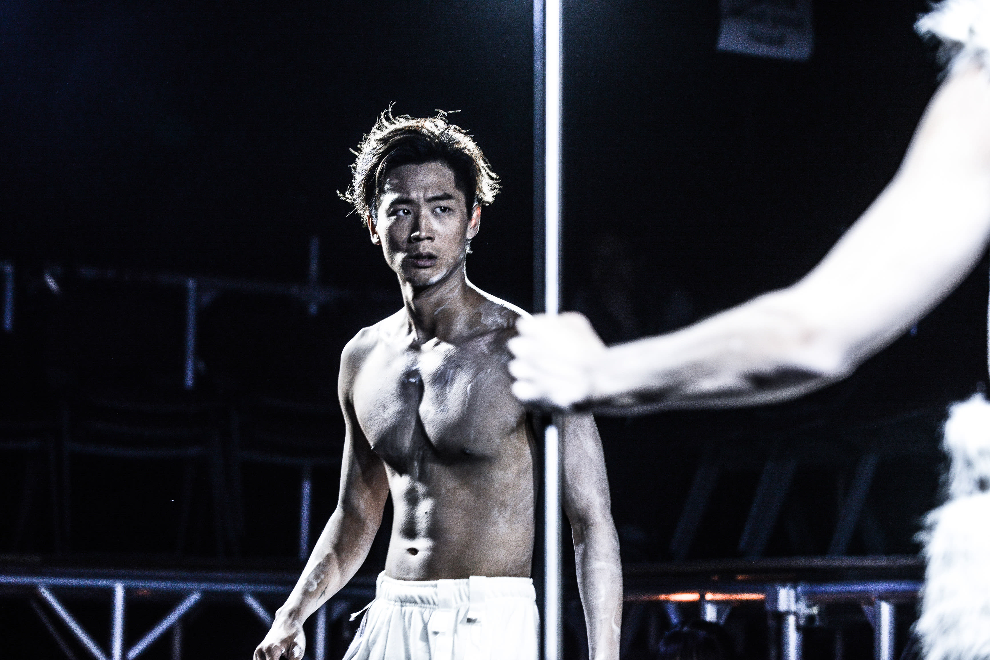 在這張來自天台製作（Rooftop Productions）當代劇場作品《行為淪喪》的劇照中，演員唐曉楓赤裸上身站在昏暗的舞台上。他的皮膚上抹著暗色妝容或污漬，呈現出這場香港舞台表演原始而感性的氛圍。他側頭凝視，表情憂慮不安，身體兩側被垂直的舞台燈光與前景中另一位表演者模糊的手臂所框住。演出名稱：《行為淪喪》