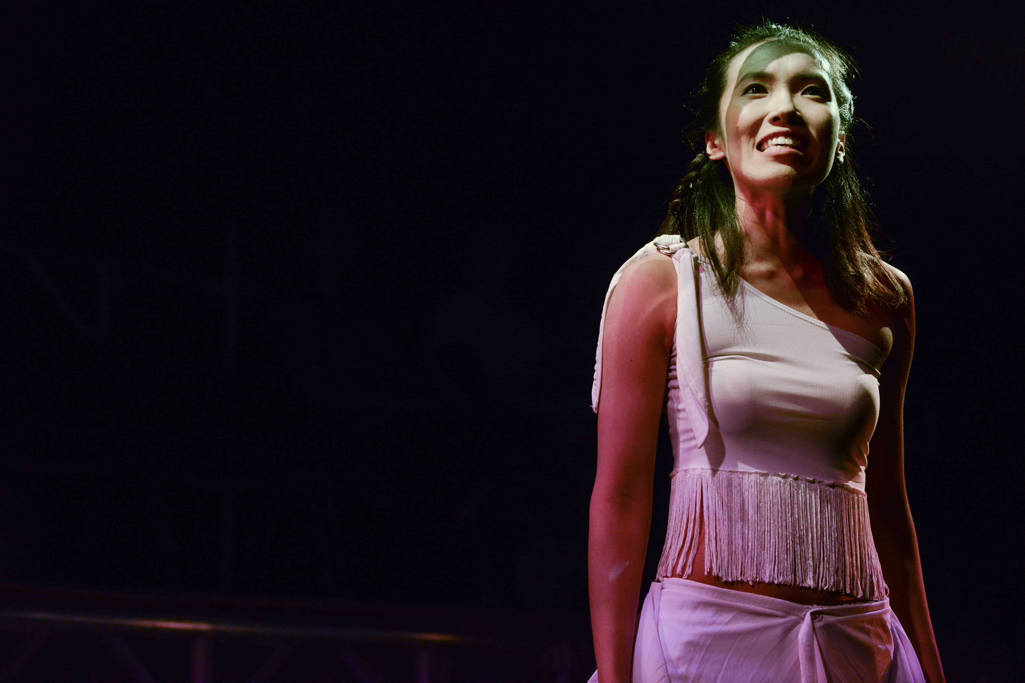 A performer stands center stage under a spotlight in a scene from the contemporary Hong Kong theatre production 'The Beautiful Ones' (行為淪喪) by Rooftop Productions. She wears a white, fringed one-shoulder top, looking upward with an expression of intense emotion. The high-contrast, dramatic stage lighting highlights her silhouette against the dark, moody background of the theatre. 'The Beautiful Ones'