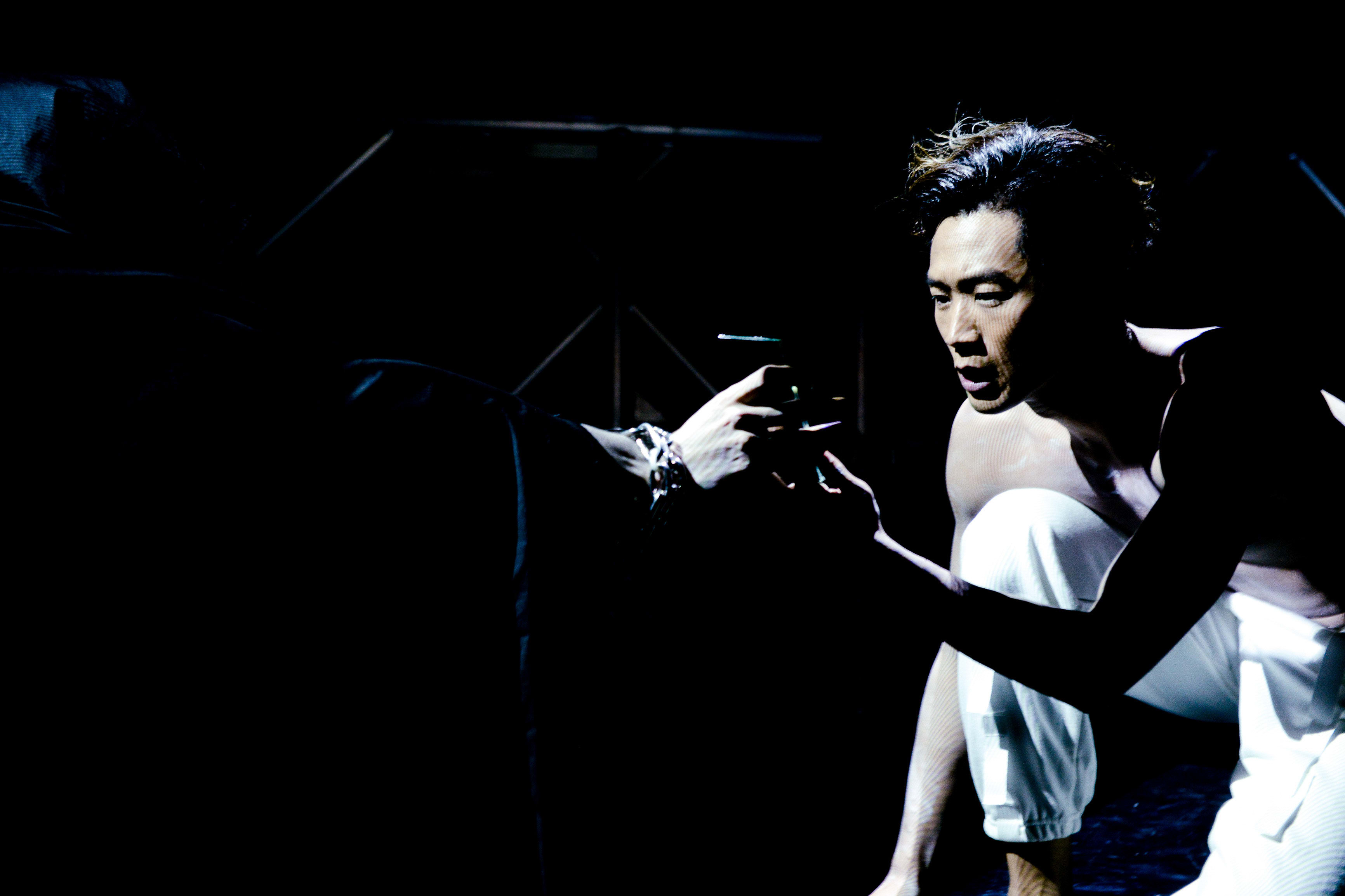 在天台製作 (Rooftop Productions) 的當代劇場作品《行為淪喪》中，演員唐曉楓身穿白色長褲，於高對比度的聚光燈下跪地。他神情凝重而脆弱，向陰影中的對手戲演員伸手，展現了該劇獨有的強烈肢體敘事張力。 「《行為淪喪》」