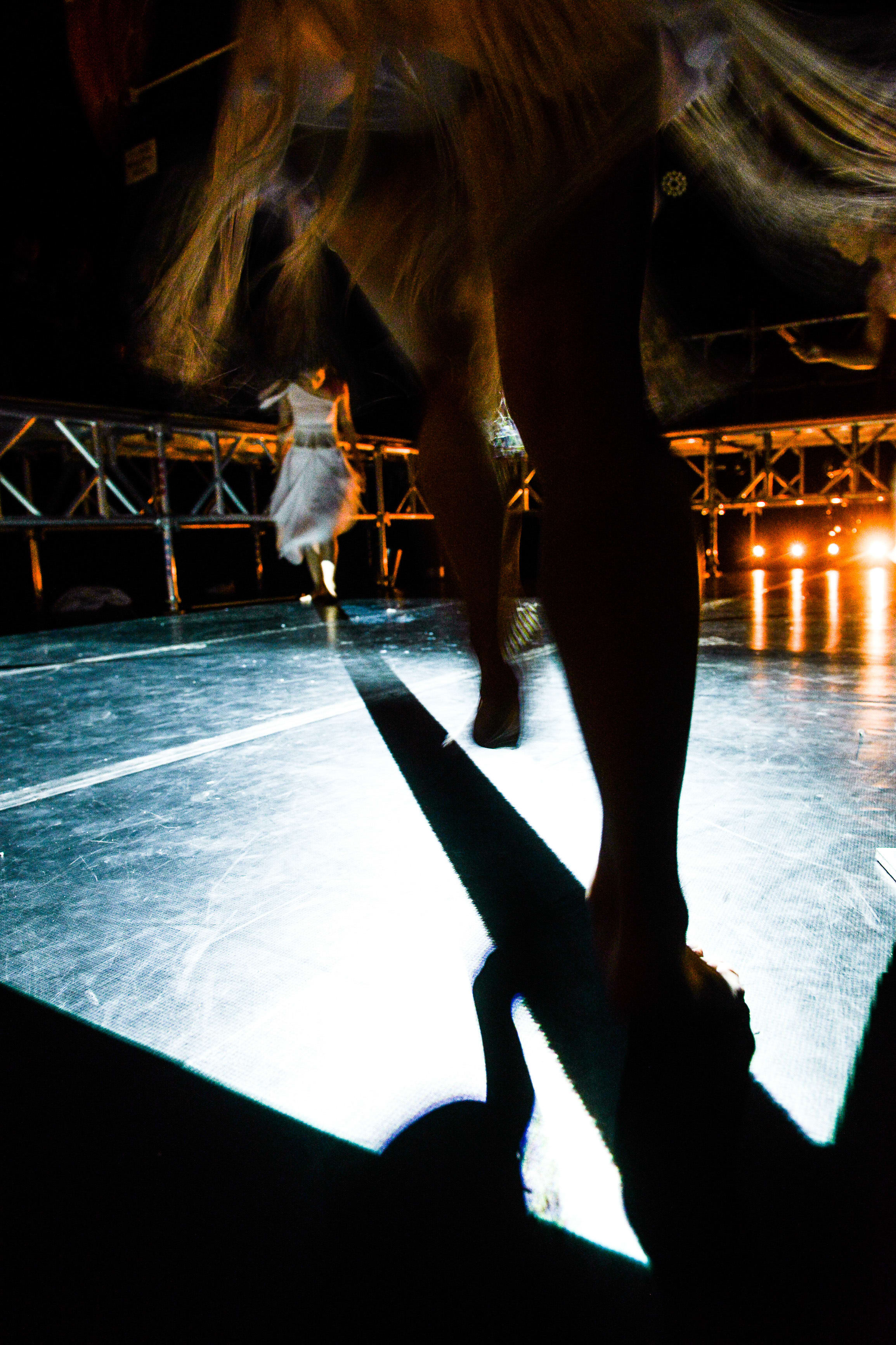 A dramatic low-angle shot from the stage floor during a contemporary theatre performance of 'The Beautiful Ones' by Hong Kong’s Rooftop Productions. In the foreground, the silhouetted legs of a performer stand on a brightly illuminated stage, casting a long, sharp shadow across the floor. In the background, another performer wearing a white dress walks away into the distance, framed by metallic scaffolding and warm stage lighting. 'The Beautiful Ones'