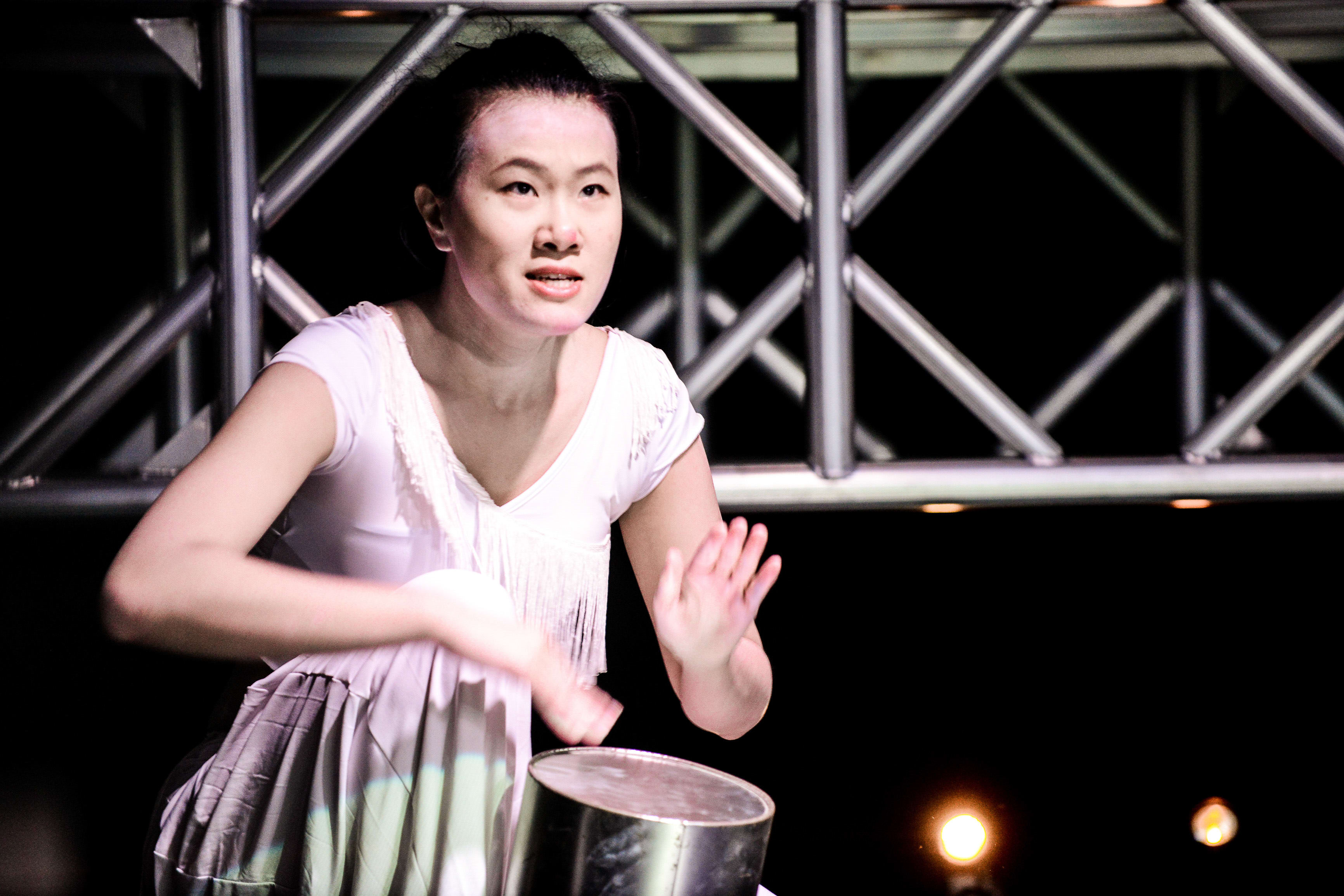 演員李婉晶正在排練天台製作（Rooftop Productions）的當代劇場作品《行為淪喪》（The Beautiful Ones）。在舞台聚光燈下，她身穿白色流蘇上衣，背景為工業風格的金屬桁架結構。李婉晶神情專注，手部正落在鼓面上，展現表演張力。《行為淪喪》
