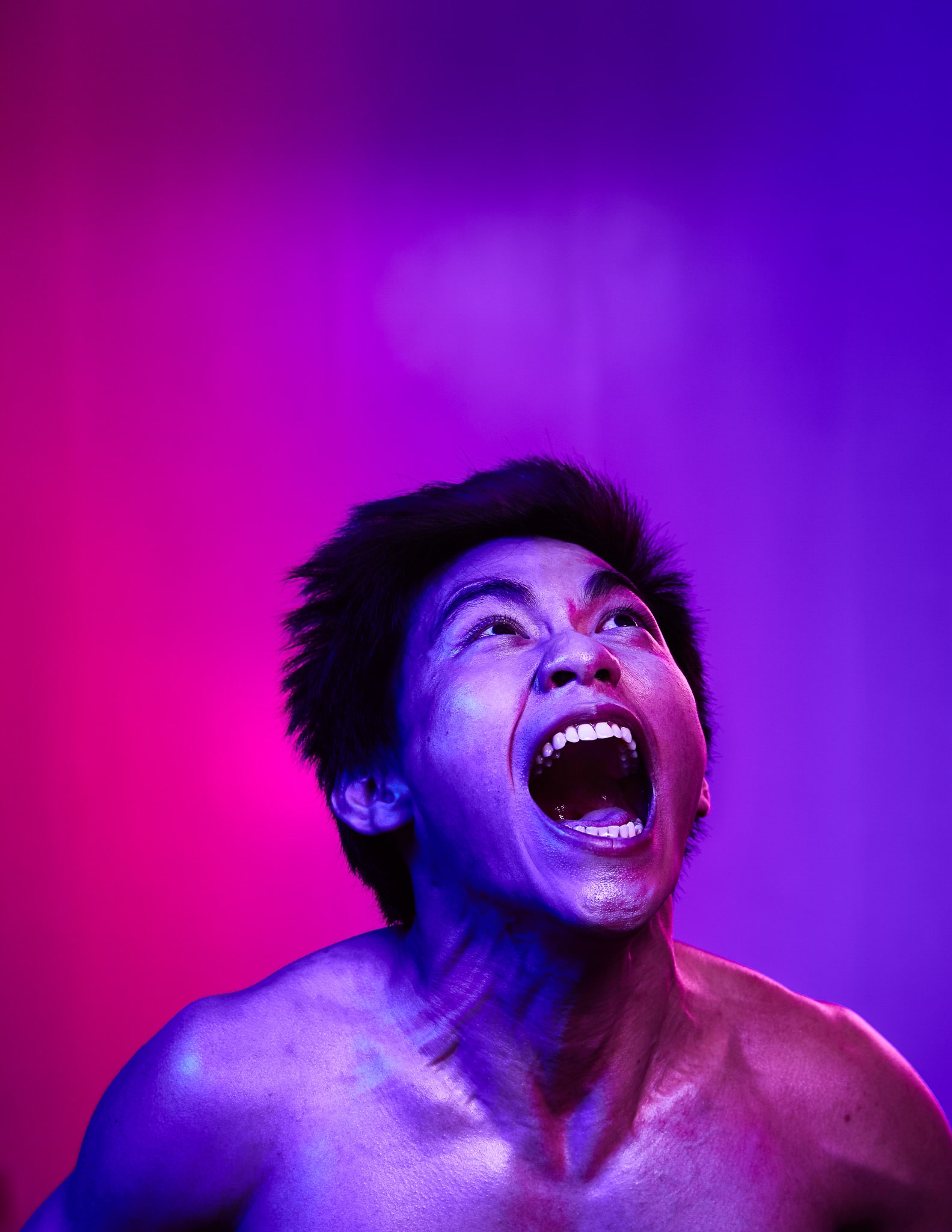 天台製作當代劇場作品《復仇變奏曲》的宣傳照，演員邱福永在強烈的燈光下進行戲劇性演繹。畫面捕捉了邱福永從胸部以上的半身像，他仰著頭，張大嘴巴，展現出極具張力與原始情感的表演瞬間。照片運用了鮮明的霓虹雙色調光影，洋紅色與深紫色在面部交織，呈現出香港舞台劇那種實驗性與感官震撼的獨特美學。邱福永為《復仇變奏曲》的宣傳照片。