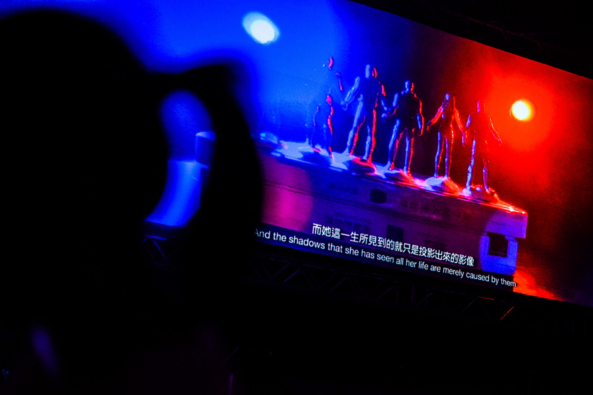 天台製作（Rooftop Productions）當代劇場作品《復仇變奏曲》的戲劇性舞台攝影。畫面呈現演員剪影佇立在潔白的高台上，在強烈對比的藍紅燈光渲染下，展現出獨特的劇場氛圍與深刻的生存隱喻，彰顯香港實驗劇場的藝術張力。影像中字幕顯示：「而佢呢一生所見到嘅就只係投影出嚟嘅影像。」