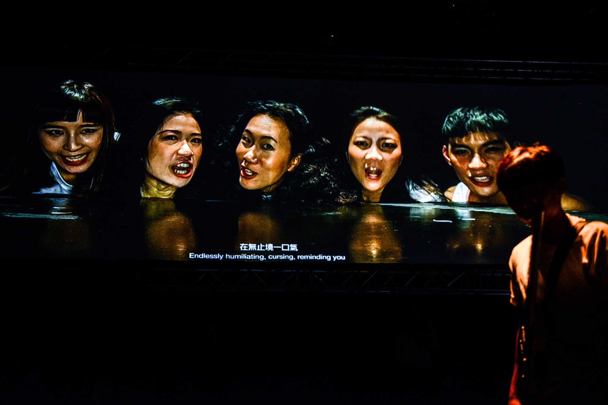 天台製作（Rooftop Productions）現代劇場作品《復仇變奏曲》（The Furies Variations）的劇照，燈光昏暗強烈，五位演員一字排開，神情猙獰激動，展現出香港實驗劇場特有的強烈張力。前景中一位表演者的剪影與舞台中央的陣容形成呼應。角色們在台上唸出：「在無止境一口氣侮辱詛咒提醒你犯下弒母的血仇。」