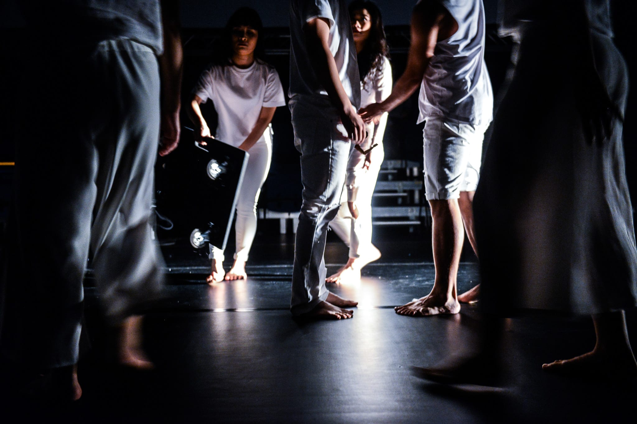 天台製作（Rooftop Productions）2018年當代劇場作品《復仇變奏曲》的舞台劇照。畫面中，演員身穿全白服裝，在幽暗簡約的舞台上進行肢體演出。隆藹宜（Lung Jes）站在後方手持錄影機，陳頴璇（Chan Wing Shuen）與其他演員在光影交錯中展現流動的當代劇場編舞，營造出濃厚的戲劇張力與神秘氛圍。香港當代劇場作品《復仇變奏曲》，天台製作，2018。