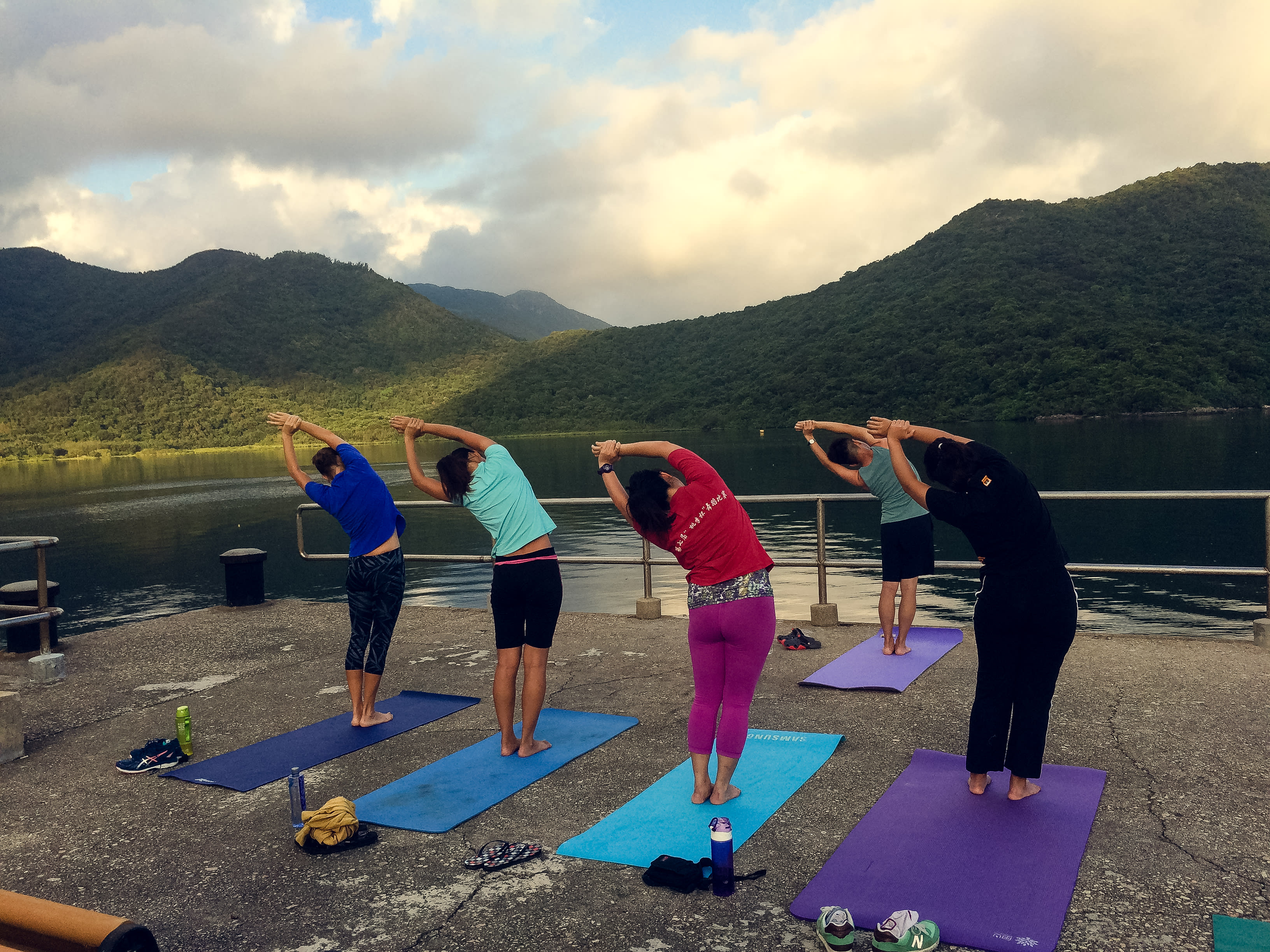 四位學員在西貢郊野公園的碼頭上練習晨間瑜伽，側身拉伸的動作融入了「當劇場走進大自然2017」（Theatre in Nature 2017）的身體劇場訓練營。在天台製作（Rooftop Productions）的帶領下，學員在平靜的湖面與蒼翠山巒的背景下，透過肢體探索與自然環境的連結。正如活動中所述：「離開城市的煩囂．．． 2017」