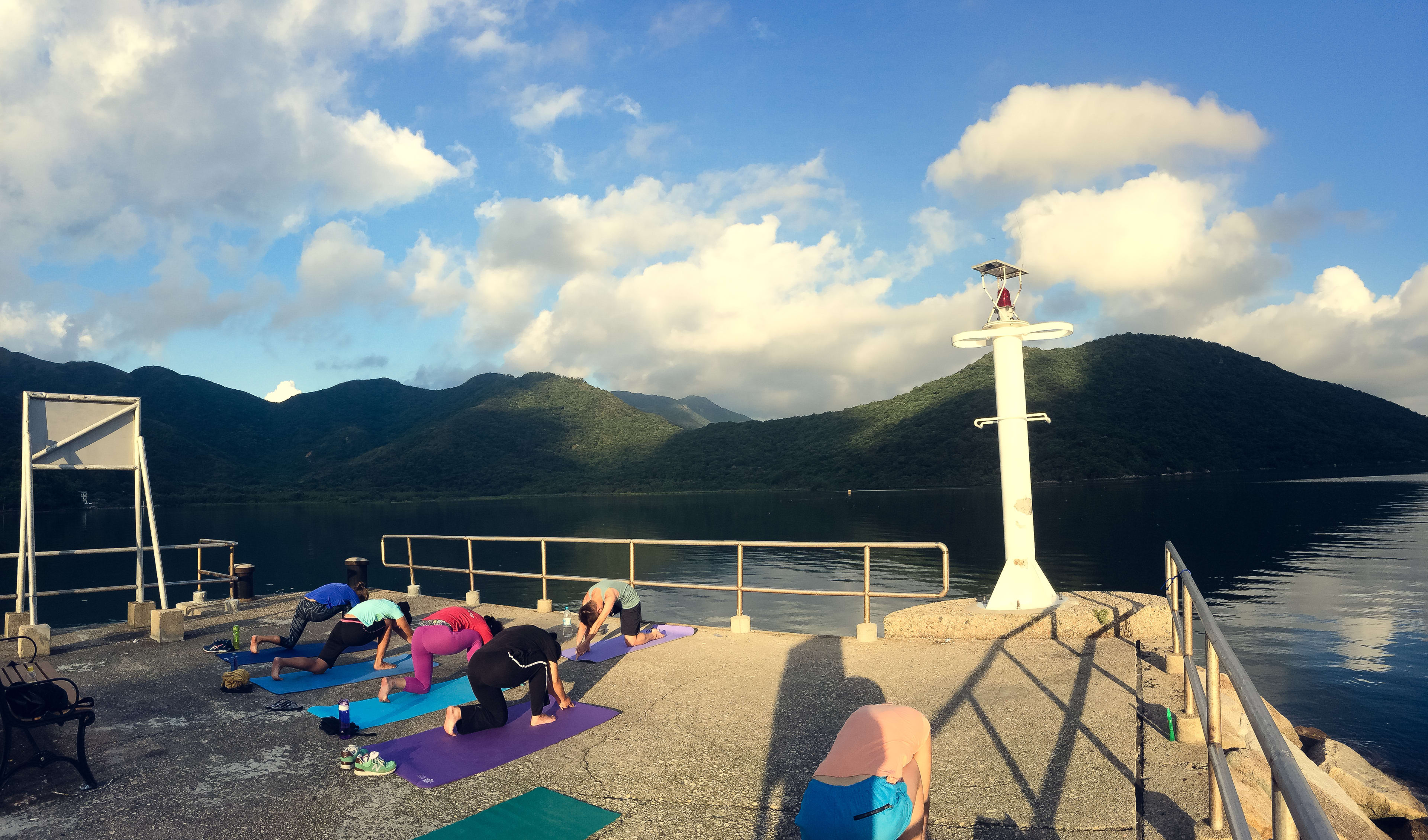 一群表演者在西貢郊野公園的一處碼頭上進行清晨瑜伽訓練。表演者在瑜伽墊上展示不同的肢體動作，背景是平靜的藍色水域和翠綠的山巒，藍天白雲下顯得格外寧靜。這是2017年「當劇場走進大自然」特定場域形體劇場訓練營的剪影，展現了香港當代劇場藝術將身體訓練與自然環境相融合的特色。「離開城市的煩囂．．． 2017」