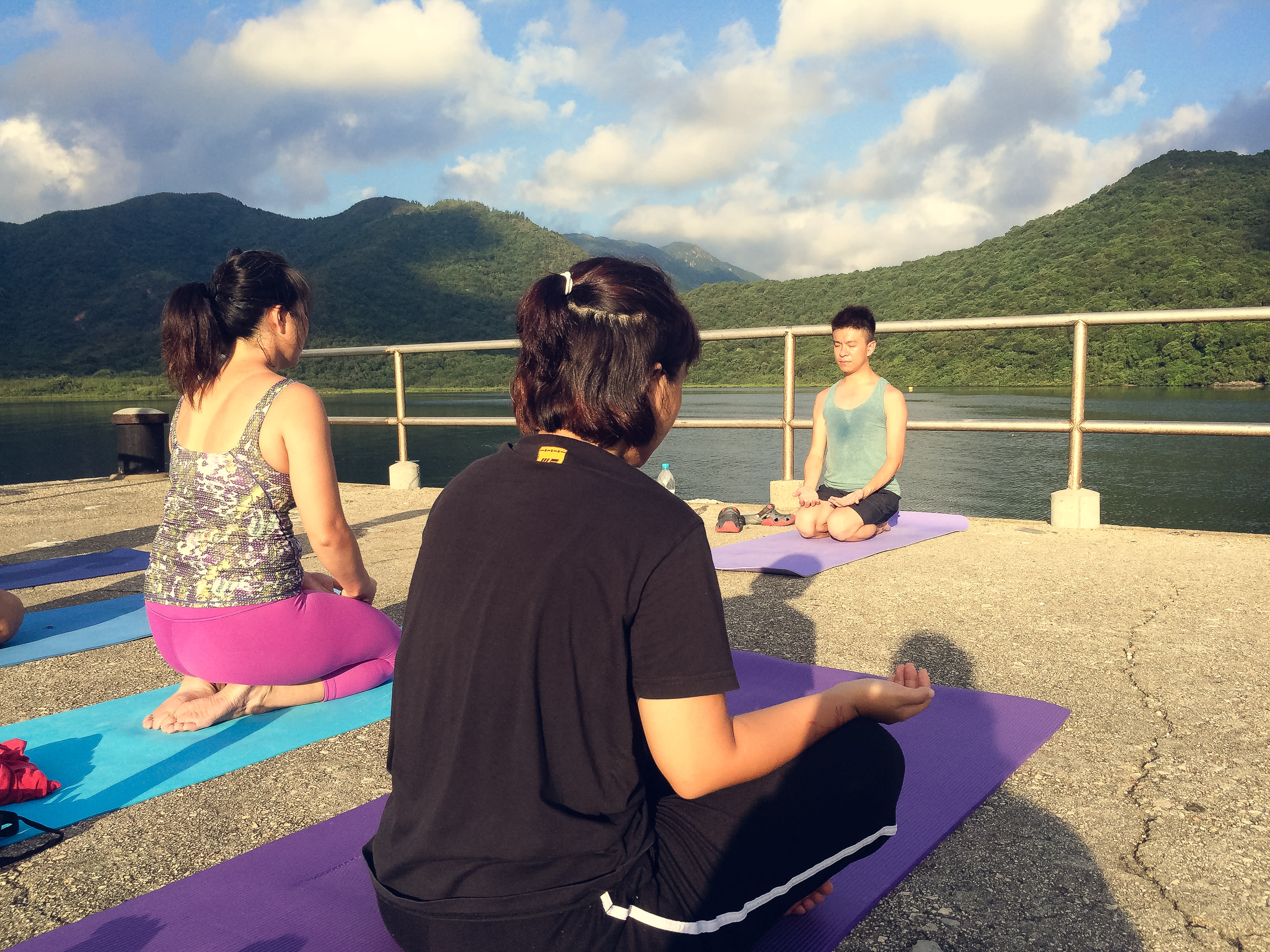 三位學員，包括施標信，正在西貢郊野公園的一處戶外平台進行「當劇場走進大自然2017」的形體與冥想訓練。在自然景致與湖泊的環繞下，參與者在金色的夕陽中練習專注與身體感知，體現了天台製作（Rooftop Productions）對於當代環境劇場的專業探索。離開城市的煩囂．．． 2017