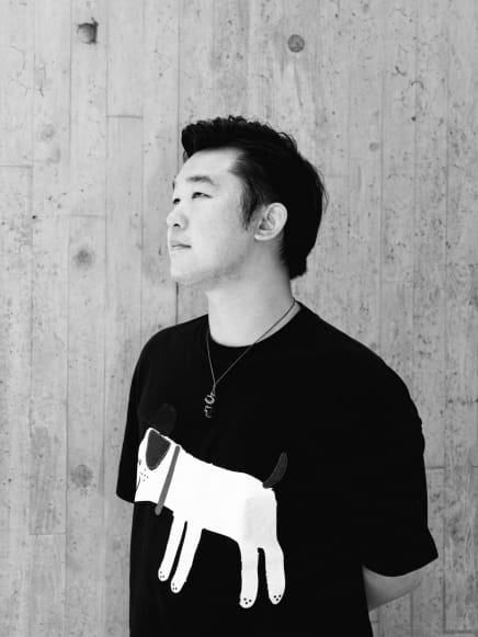 一張邱永亮（Thomas Yau）的黑白定裝照，背景為充滿質感的垂直條紋混凝土牆。照片中，邱永亮身穿印有狗圖案的黑色T恤，佩戴著精緻的項鍊，側身站立，神情冷靜而深思，展現出天台製作（Rooftop Productions）當代香港劇場表演中細膩的情感氛圍。定裝照。
