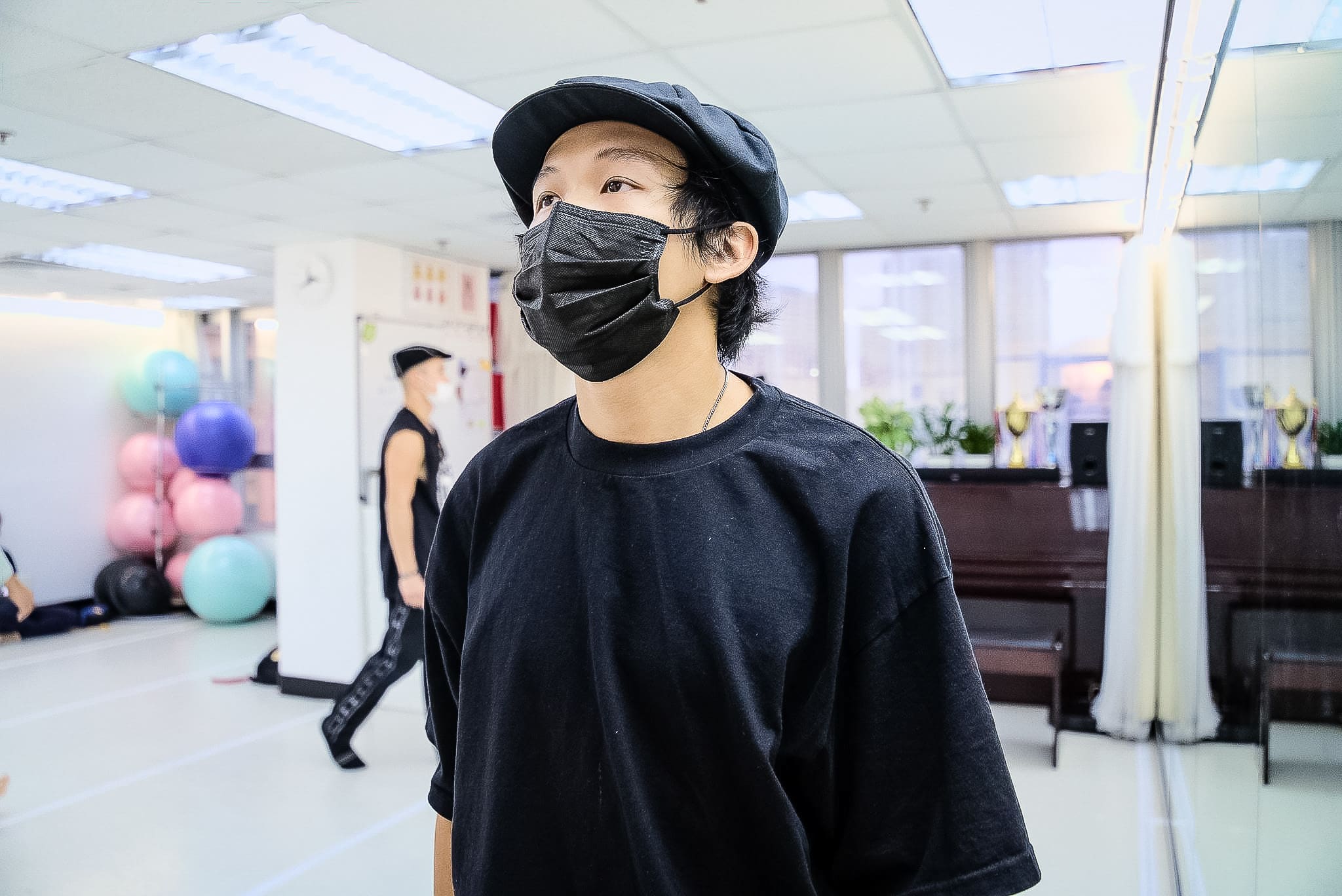 在天台製作（Rooftop Productions）當代劇場作品《舞照跳》的排練室中，演員廖偉傑（New Y-Kid）頭戴黑色報童帽並佩戴口罩，若有所思地望向側方。背景中另一位表演者走過擺滿彩色瑜伽球的排練場，展現了這部香港舞台劇的動態排練氛圍。表演者沉浸在對工業時代的回憶中說道：「六十年代那時有一大堆工廠。」