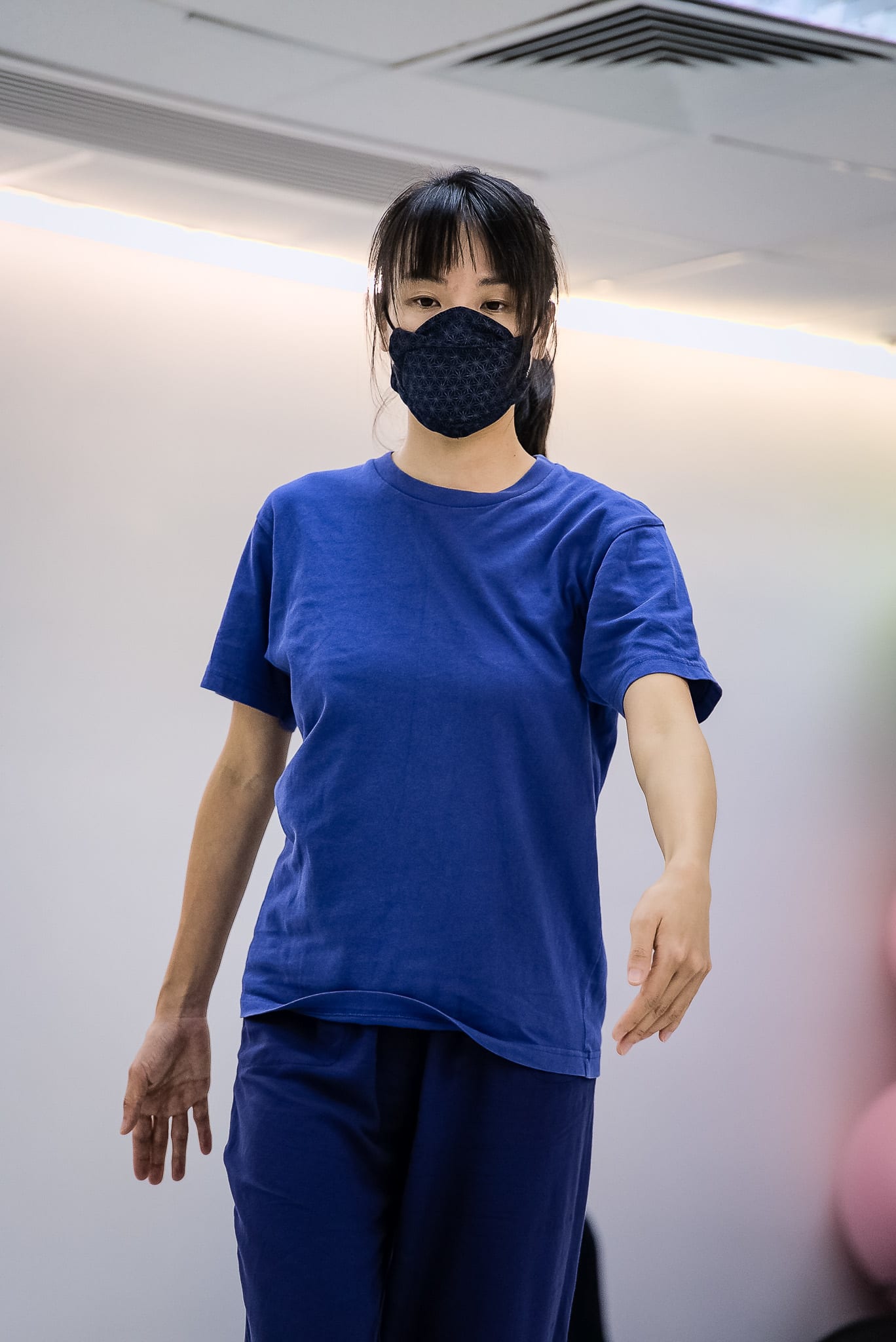 一名舞者在香港當代劇場製作《舞照跳》（We Still Dance）的排練中穿過排練室舞台。她身穿簡約的藍色T恤、深色長褲並配戴花紋口罩，神情專注、姿態堅定，展現了當代舞蹈劇場的誠摯張力。在移動的過程中，她述說著：「五十至七十年代，城市發展。」