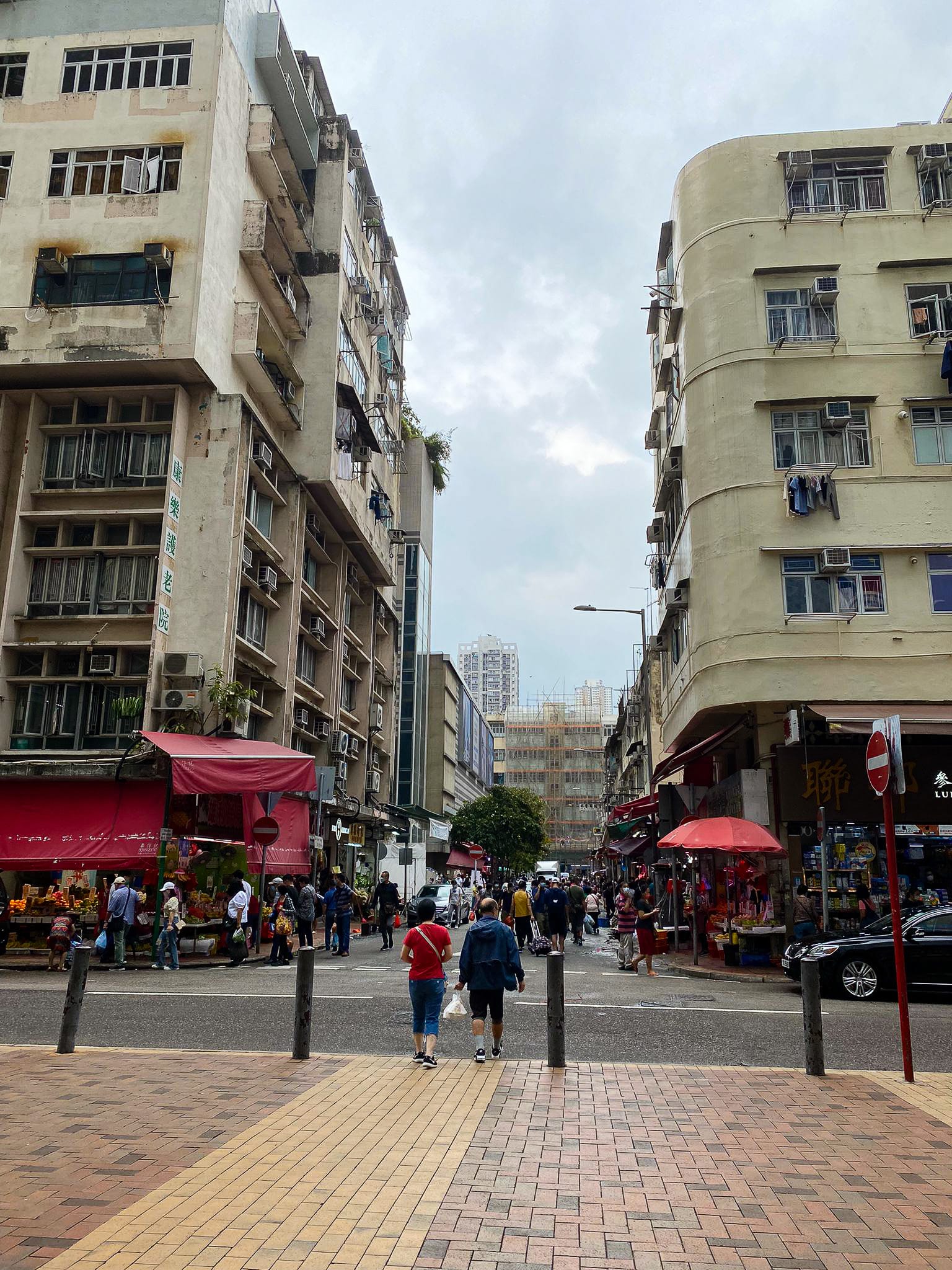 從街道視角拍攝的香港窄巷，兩旁林立著充滿歲月痕跡的住宅大廈，為天台製作的當代劇場作品《舞照跳》營造出充滿電影感的場景。行人正穿過繁忙的十字路口，陰天的光影交織出日常生活的真實律動，呼應著演出中關於城市與記憶的題旨。「消逝。」