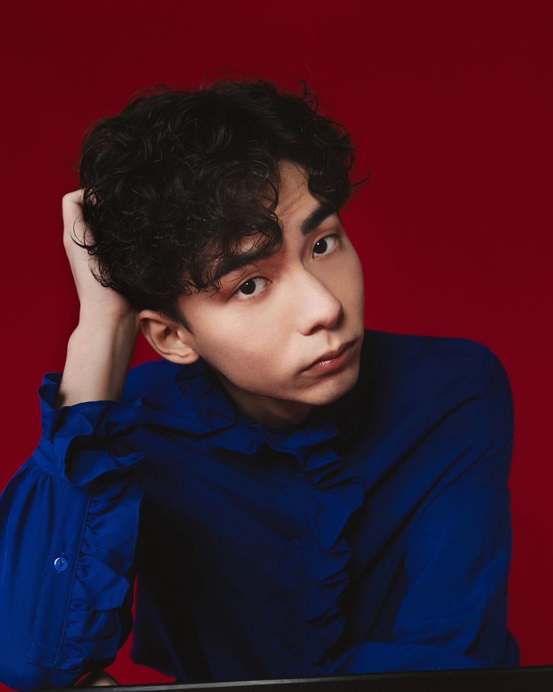 A studio headshot of Hong Kong actor Wu Lui-fung for a Rooftop Productions (天台製作) theatrical performance. He is wearing a long-sleeved navy blue ruffled shirt and posing with one hand resting behind his head, looking directly at the camera with a thoughtful, contemplative expression against a solid, deep red background. The dramatic lighting emphasizes his textured, dark curly hair and the minimalist aesthetic characteristic of contemporary HK theatre.