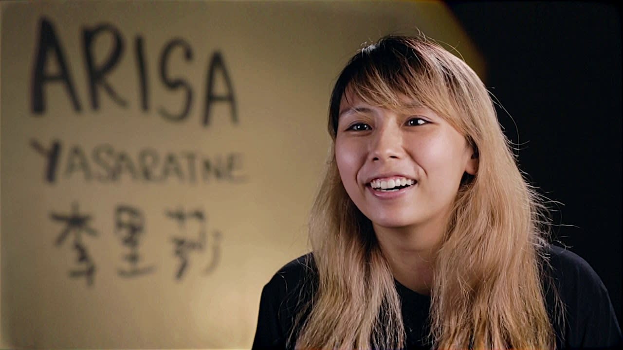 天台製作最新舞台劇《山下的證詞》宣傳影片預覽圖，以電影感寬銀幕比例呈現。畫面中演員李里莎（Arisa Yasarane）於溫暖的室內環境中，對著鏡頭展露親切自然的笑容。背景牆上寫有「ARISA YASARATNE」及其中文名字「李里莎」的手寫字樣，展現出這場香港當代劇場演出背後，演員真摯的一面。