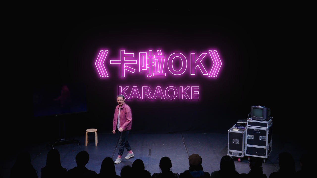 天台製作當代劇場作品《卡啦OK》演出精華宣傳影片縮圖，採用 16:9 寬銀幕構圖。畫面呈現一個極簡的黑盒劇場空間，身穿粉紅色外套的演員正走過舞台前往觀眾席方向。背景上方懸掛著顯眼的粉紅色霓虹燈招牌，寫著《卡啦OK》與英文名「KARAOKE」。舞台右側放置了一台復古電視機及專業演出器材箱，展現了香港前衛劇場獨有的演出美學與製作細節。