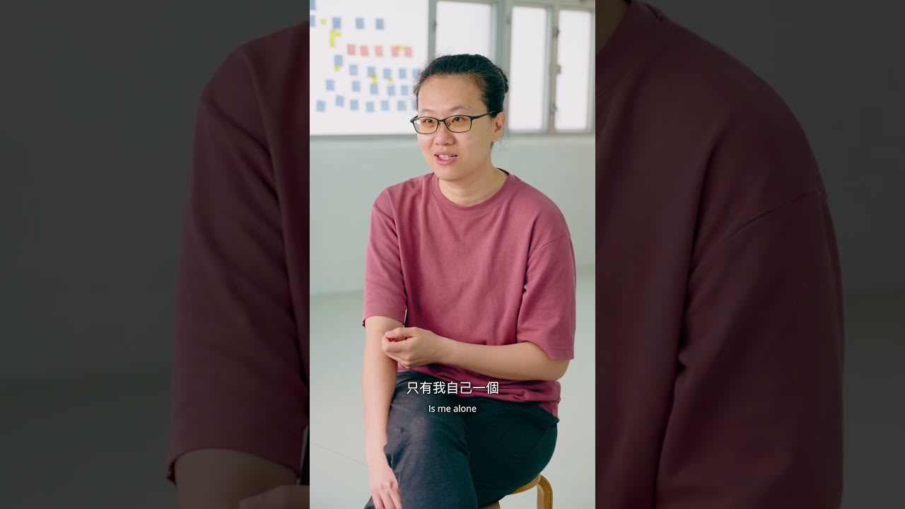 天台製作（Rooftop Productions）紀錄片《卡啦OK》的直式影片縮圖，展示了香港劇場創作者李婉晶（Michelle Li）在明亮工作室中的特寫。她身穿棗紅色上衣，戴著眼鏡，神情專注且若有所思地看向鏡頭。背景隱約可見貼滿彩色便條紙的白板，體現了當代香港劇場創作的排練過程。影像風格簡約自然，充滿藝文氣息。縮圖內含引述：「在香港劇場界走過十一年，《卡啦OK》是她首次推出的個人作品。」