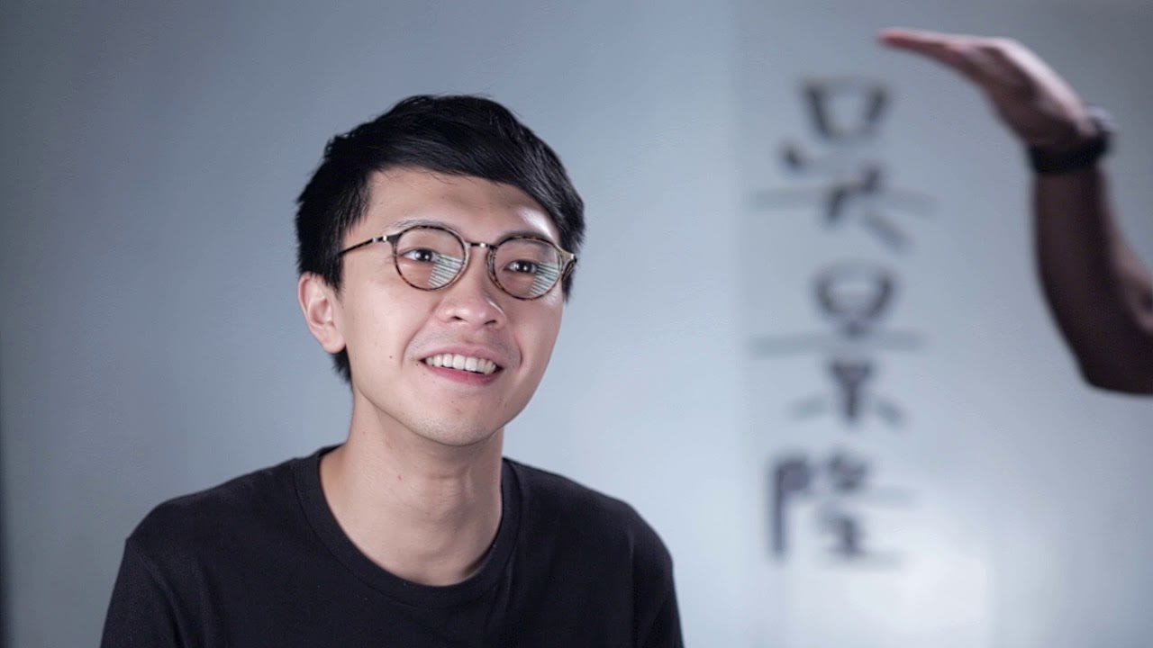 《山下的證詞》舞台劇宣傳短片縮圖，展示演員吳景隆的深度訪談。此寬螢幕影像以中景鏡頭捕捉吳景隆的半身像，背景為柔和的影棚光線。他佩戴黑框眼鏡並身穿黑色上衣，神情輕鬆自然，正對著鏡頭侃侃而談。右側背景隱約可見該劇目的書法題字，一隻手部動作出現在畫面邊緣，突顯這部當代香港劇場作品的幕後創作氛圍。
