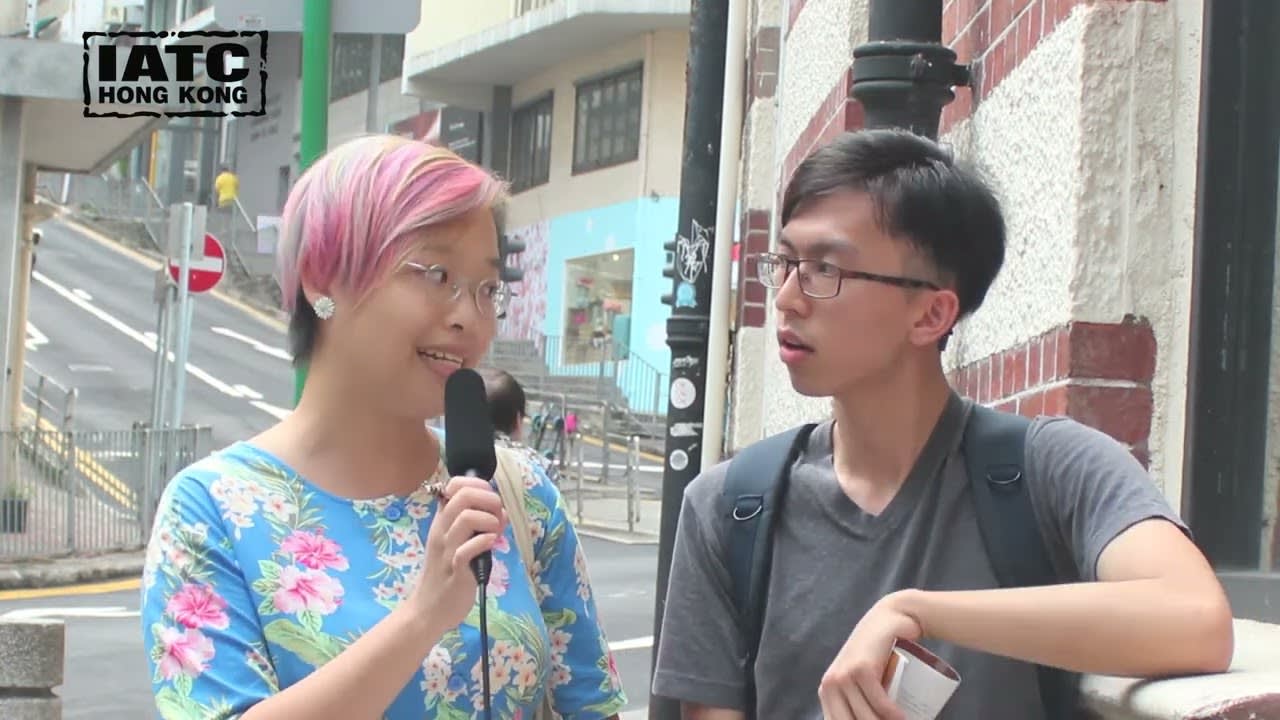天台製作《牛奶與蜜糖》的錄影劇評截圖，呈現香港街頭訪問的場景。畫面中，留著粉色挑染短髮的女記者穿著藍色花紋上衣手持麥克風，正在訪問一位身穿灰衣的青年，對方正專注聆聽。背景為香港常見的斜坡街道與城市景觀，捕捉了該現代劇場作品質樸、充滿能量的演出風格。圖片附帶引言：「這個團隊在選擇道具方面，意象非常精準，當中富有幽默感，亦扣連到遷徙的主題。我會欣賞整個風格，質樸之餘，能量亦充沛；整體感覺非常清新及流暢。」