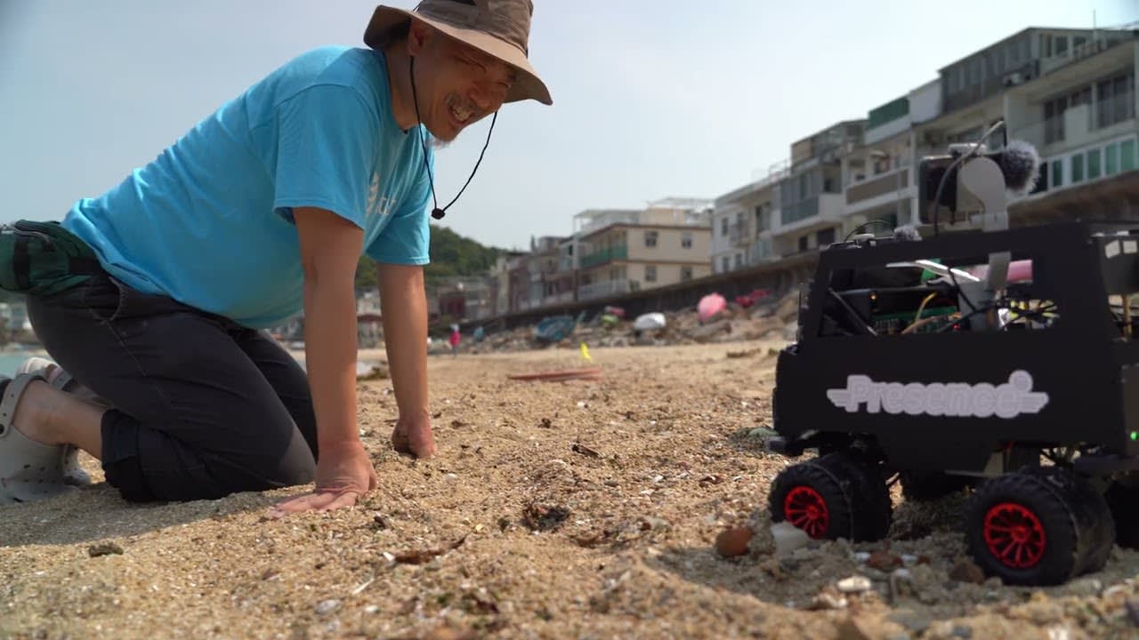 Promotional video thumbnail for the contemporary Hong Kong theatre production 'Presence' (遙距在場), featuring a widescreen cinematic shot on a sandy beach. A man wearing a bucket hat and a bright blue t-shirt is kneeling in the sand, smiling warmly as he interacts with a small, remote-controlled robot labeled 'Presence'. The setting suggests a blend of technology and human connection, characteristic of modern experimental stage performance. The scene is brightly lit with a hazy, natural outdoor aesthetic, capturing a moment of discovery.