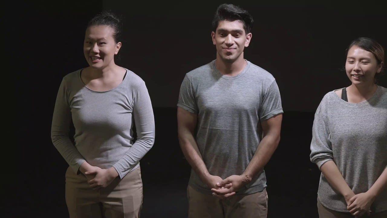 天台製作（Rooftop Productions）劇目《山下的證詞》完整演出視頻的寬螢幕劇照縮圖。影像展示三名演員並排站立在昏暗、極簡的舞台上。中間的男演員身穿灰色T恤，兩側分別站著穿著中性灰色調休閒上衣的女演員，眾人神情平靜而沉思，面向觀眾。戲劇性的低調燈光將他們的面部表情從黑暗中襯托出來，突顯出這部香港當代劇場作品原始而親密的特質。