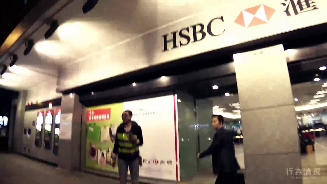 天台製作當代劇場作品《行為淪喪》音頻導覽的寬銀幕視頻縮圖。畫面呈現香港匯豐銀行分行外的夜景，燈光昏暗並帶有戲劇性的模糊動態，捕捉到兩名途人經過銀行玻璃門面的瞬間。構圖體現了該劇作場域特定（site-specific）與沉浸式的表演風格，展現出香港獨立劇場的獨特氣質。