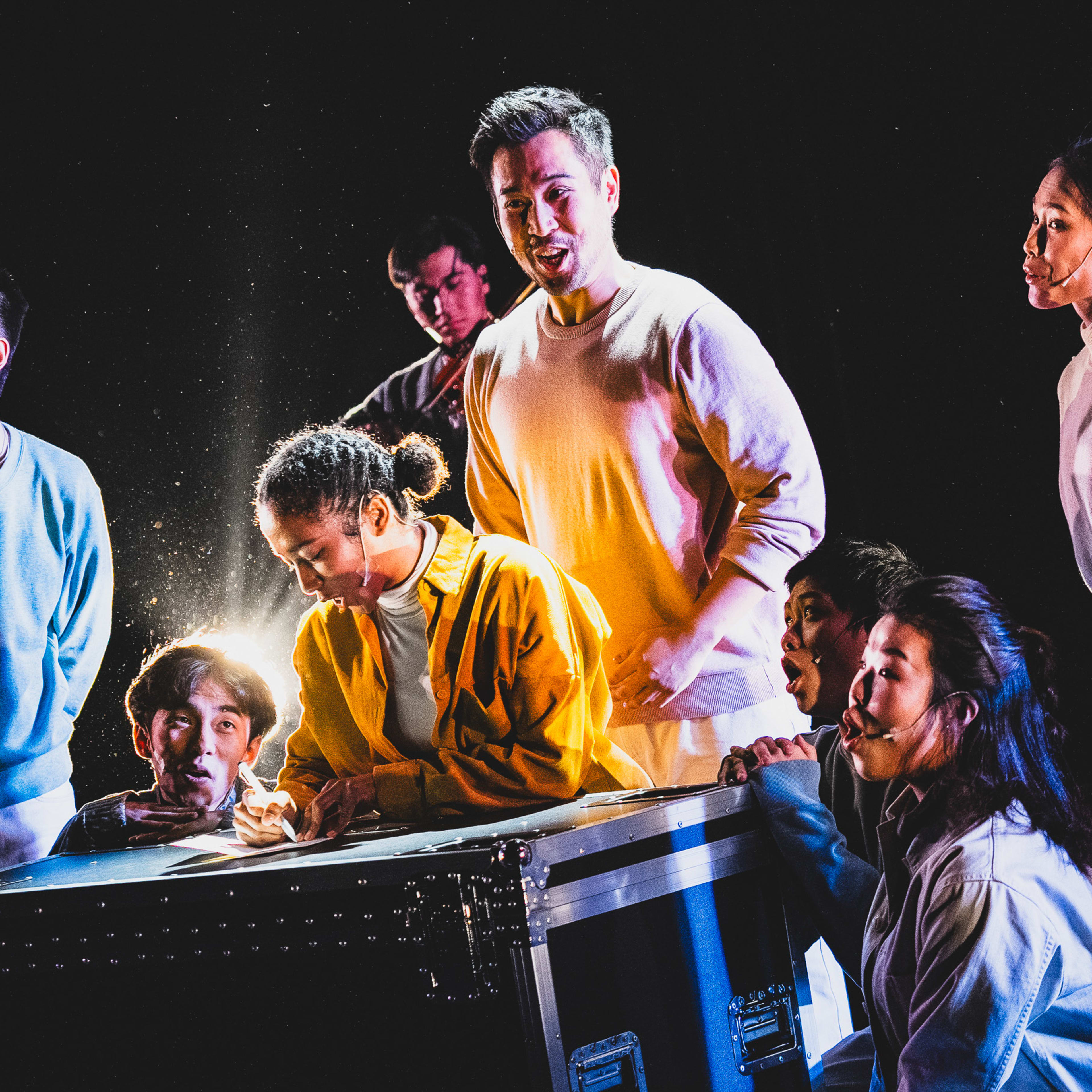 天台製作（Rooftop Productions）當代劇場作品《天真與世故之歌》的舞台演出劇照。在強烈且戲劇性的燈光下，演員們圍聚在一個黑色大型器材箱旁。身穿亮黃色外套的演員正低頭專注地在箱上書寫，聚光燈在該處營造出焦點，而周圍的演員（包括莫珏邦、陳琬瑜等）神情各異，展現出緊張與期待的氛圍。這場香港當代劇場演出融合了實驗性表演與極具張力的舞台調度。《天真與世故之歌》（西九）
