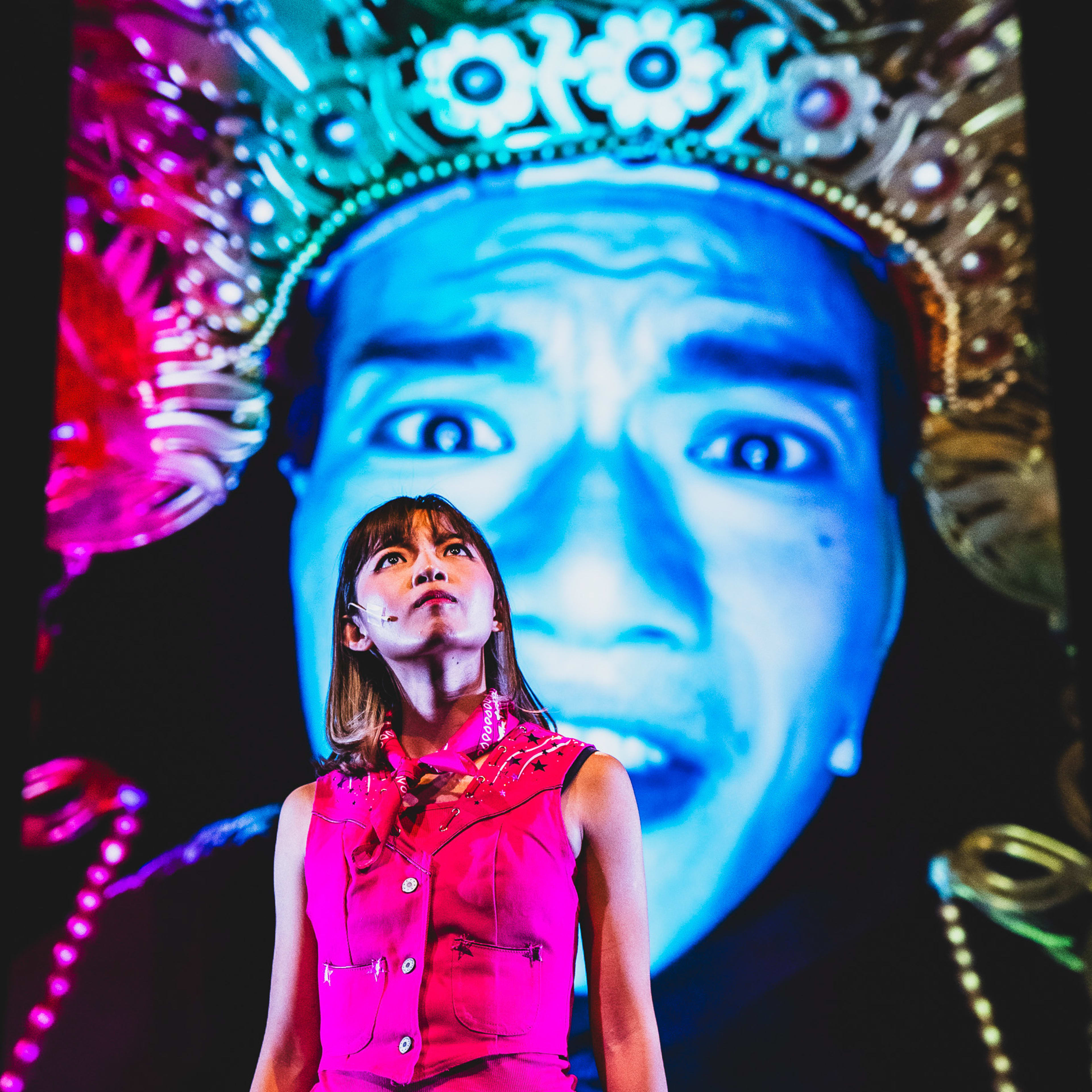 在香港當代劇場製作《天真與世故之歌》(Songs of Innocence and Experience) 的西九演出中，演員冼樂欣身穿鮮粉紅色無袖背心與短裙，站在舞台中央。她神情堅定地向上仰望，舞台燈光戲劇性地投射在她身上。在她身後，巨大的數碼投影呈現出羅文澤戴著精緻傳統風格頭飾的面部特寫，為表演增添了強烈而超現實的視覺張力。《天真與世故之歌》（西九）