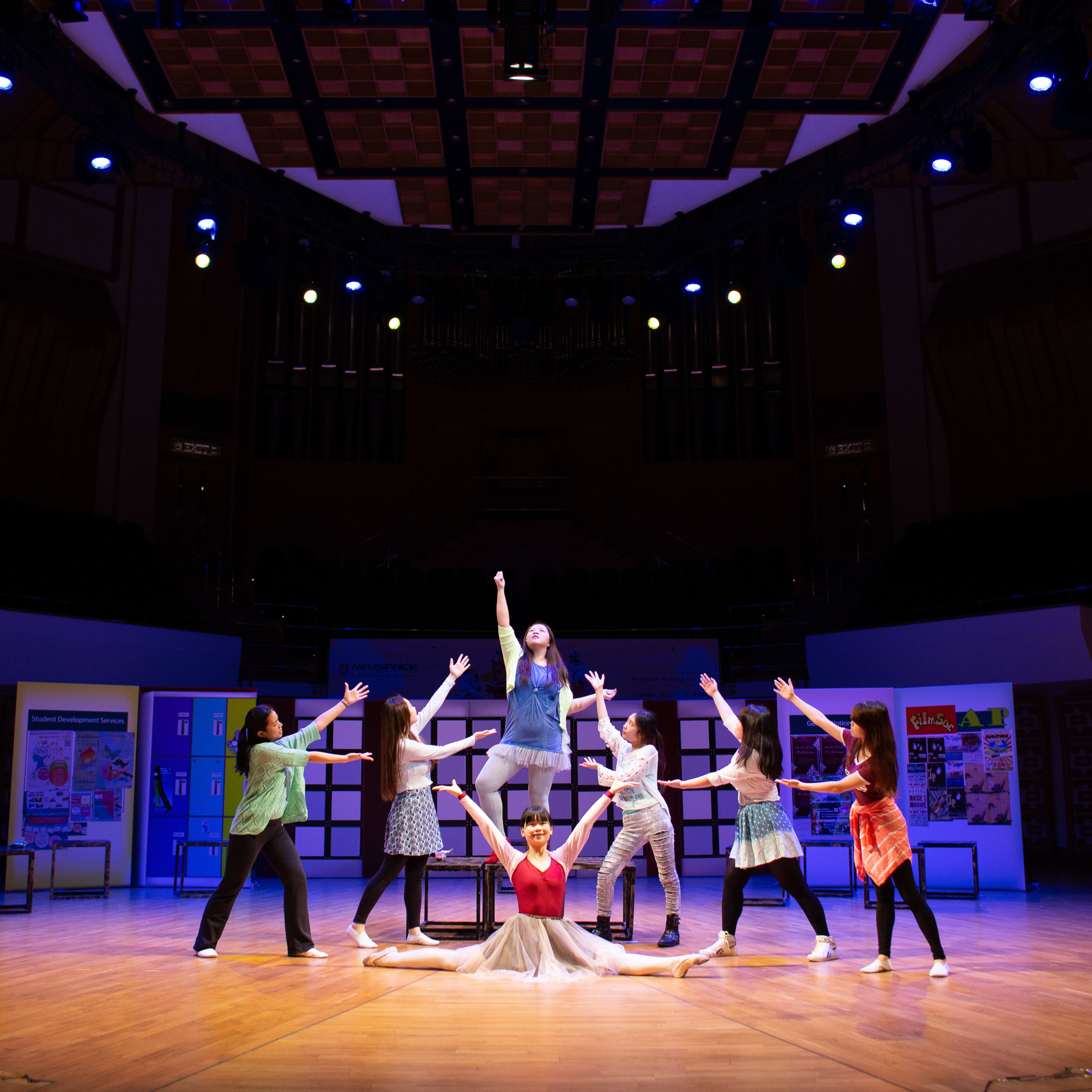 香港天台製作 (Rooftop Productions) 的一群舞者在專業劇場舞台上，為音樂劇《Fame》進行精彩演出。畫面中央的表演者展現出色的側劈腿動作，雙手伸向空中，其餘六名演員圍繞在旁，動作各異，充滿了慶祝與專注的神采。柔和的舞台聚光燈映照在表演者身上，與幽暗的禮堂形成鮮明對比，呈現出當代劇場表演的活力與熱情。其中一位演員深情唱道：「Fame」。