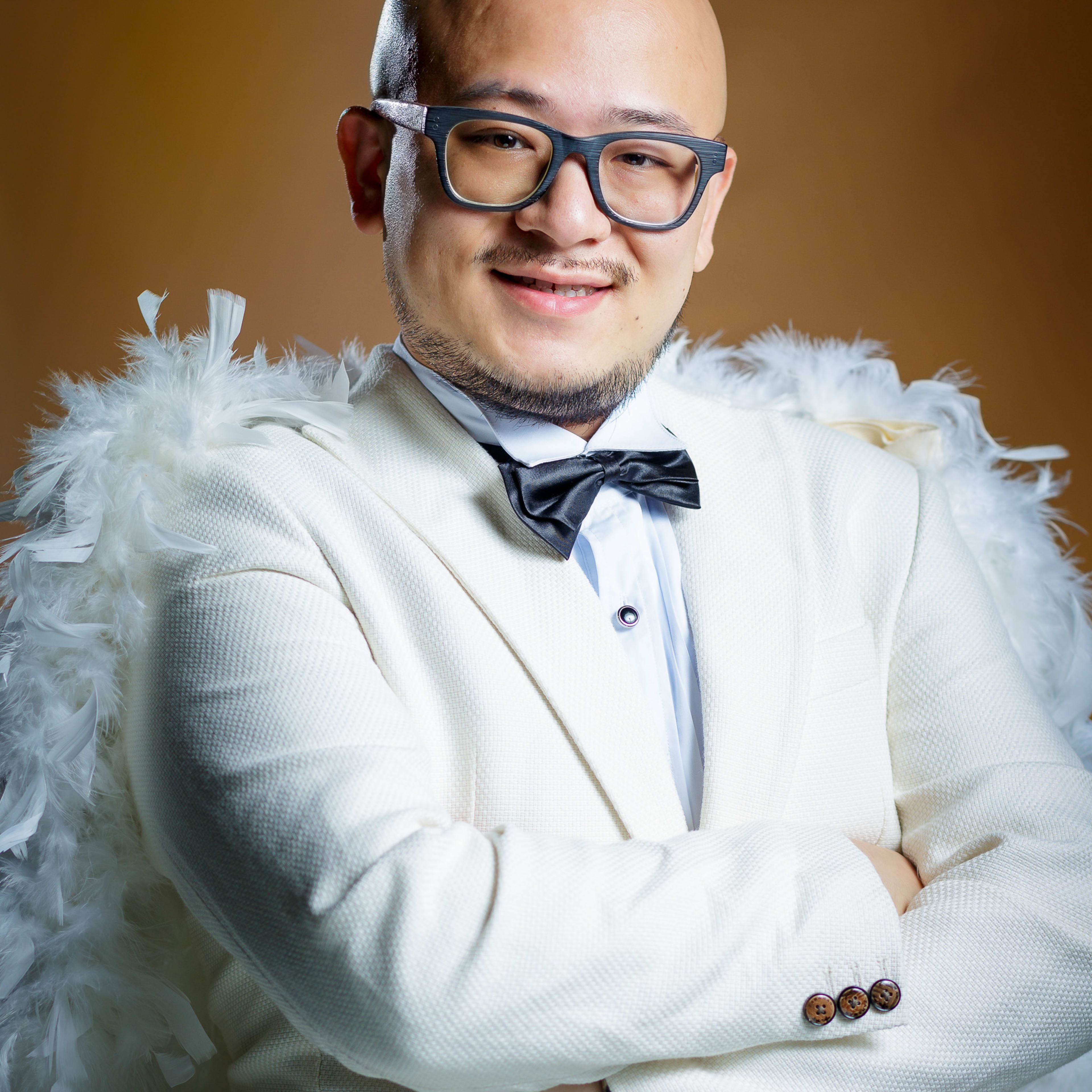 Portrait of performer Alfie Leung for the Rooftop Productions contemporary Hong Kong theatre show 'Superheroes Don't Give a Shit'. Leung is wearing a white tuxedo jacket paired with a white feather boa, styled as a whimsical, satirical take on a superhero costume. He is captured in a studio setting with warm professional lighting, posing with his arms crossed and a gentle, inviting smile. 'Superheroes Don't Give a Shit!'