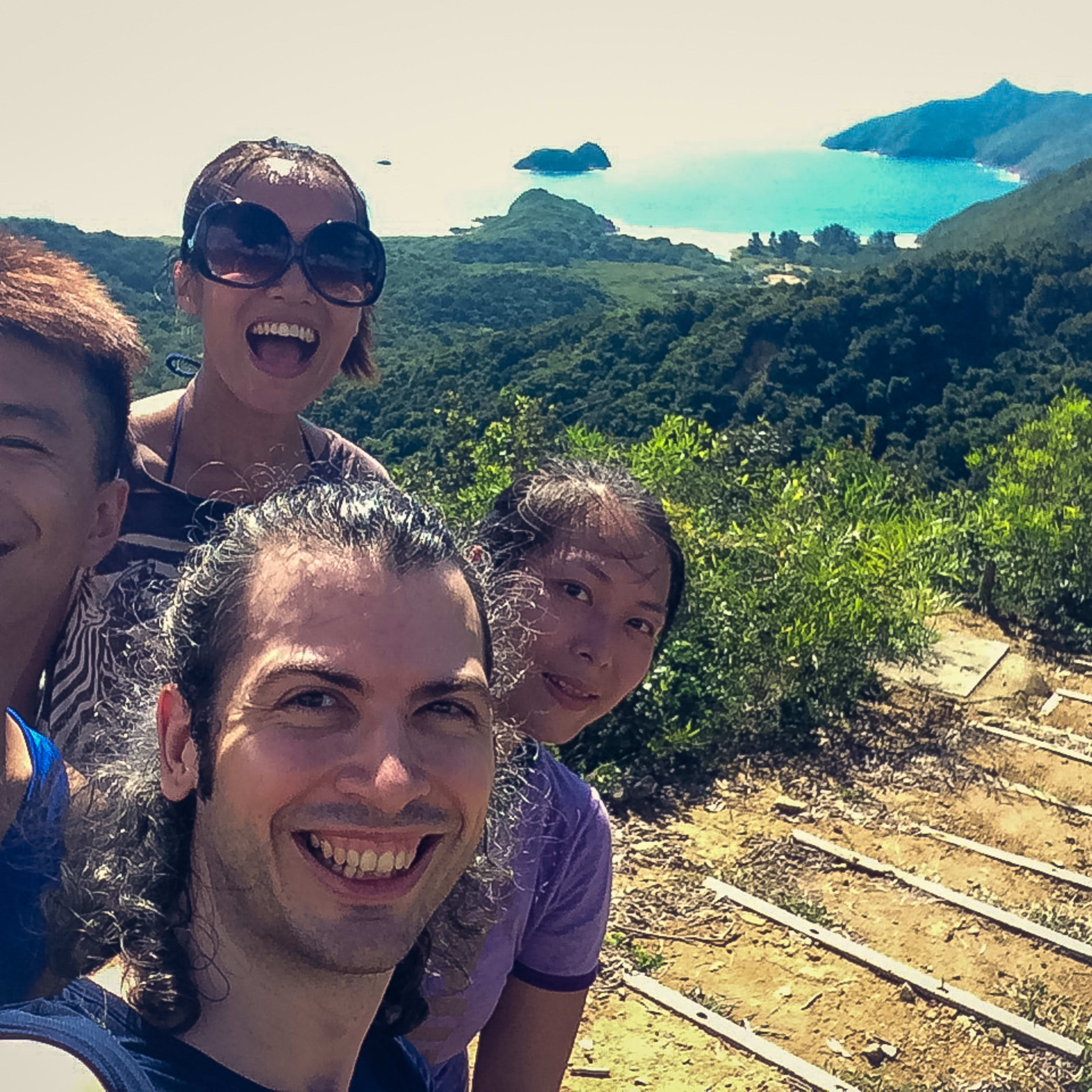 A vibrant selfie of actors Billy Sy and Ivor Houlker alongside colleagues during a 'Theatre in Nature 2017' physical theatre workshop in Sai Kung Country Park. The group is captured hiking on a sun-drenched trail overlooking the coastline, showcasing the intersection of contemporary Hong Kong site-specific performance and the great outdoors. 'Theatre in Nature, a site-specific physical theatre training camp in Sai Kung Country Park - hiking'