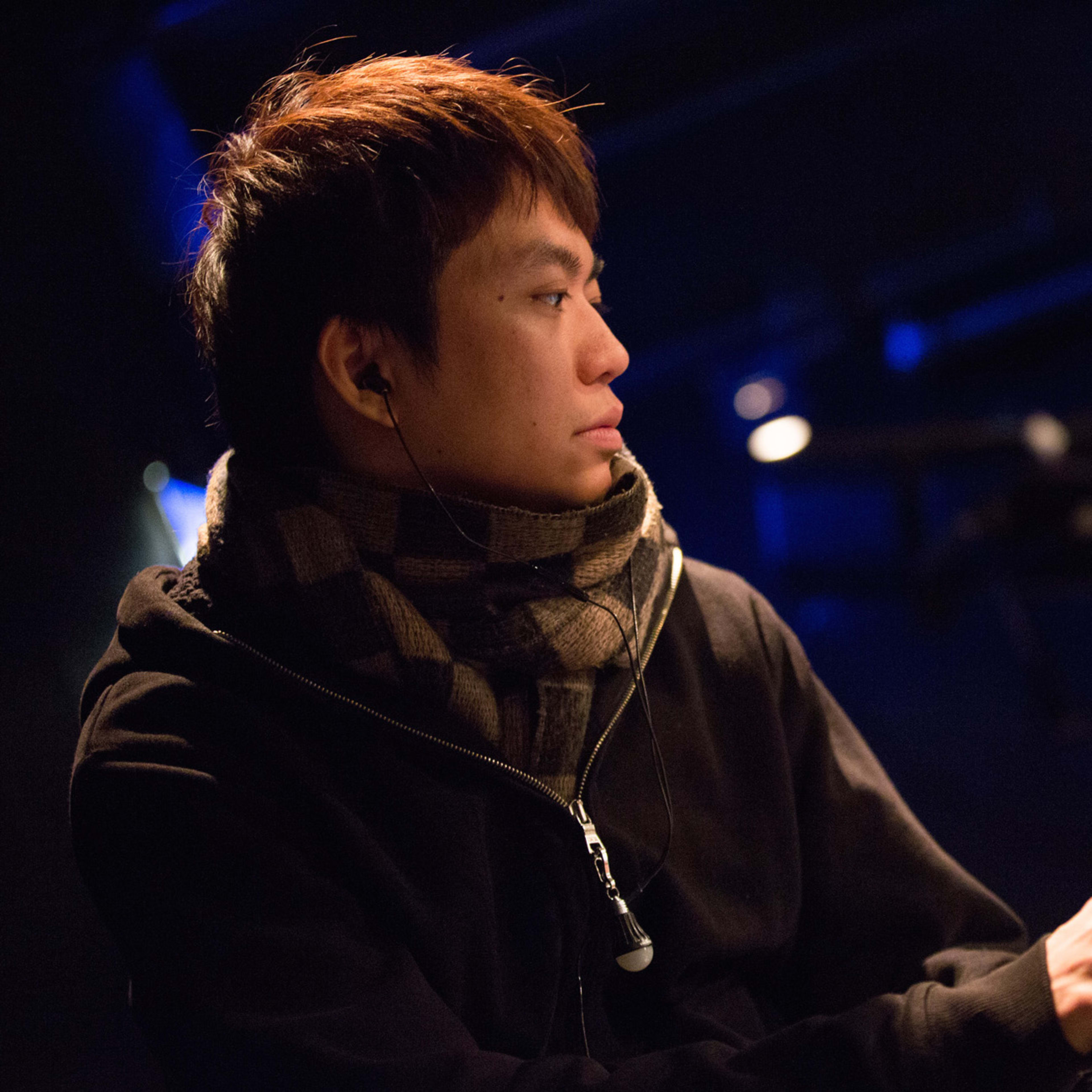 歐陽翰奇（Au Yeung Hon Ki）於天台製作（Rooftop Productions）當代香港劇場排練期間的側拍特寫。照片中他身穿深色連帽衫與格紋圍巾，正專注地注視著舞台控制台上的筆記型電腦螢幕，周圍環繞著劇場特有的冷調藍光。這張照片捕捉了演出前的技術排練時刻。個人特寫照。