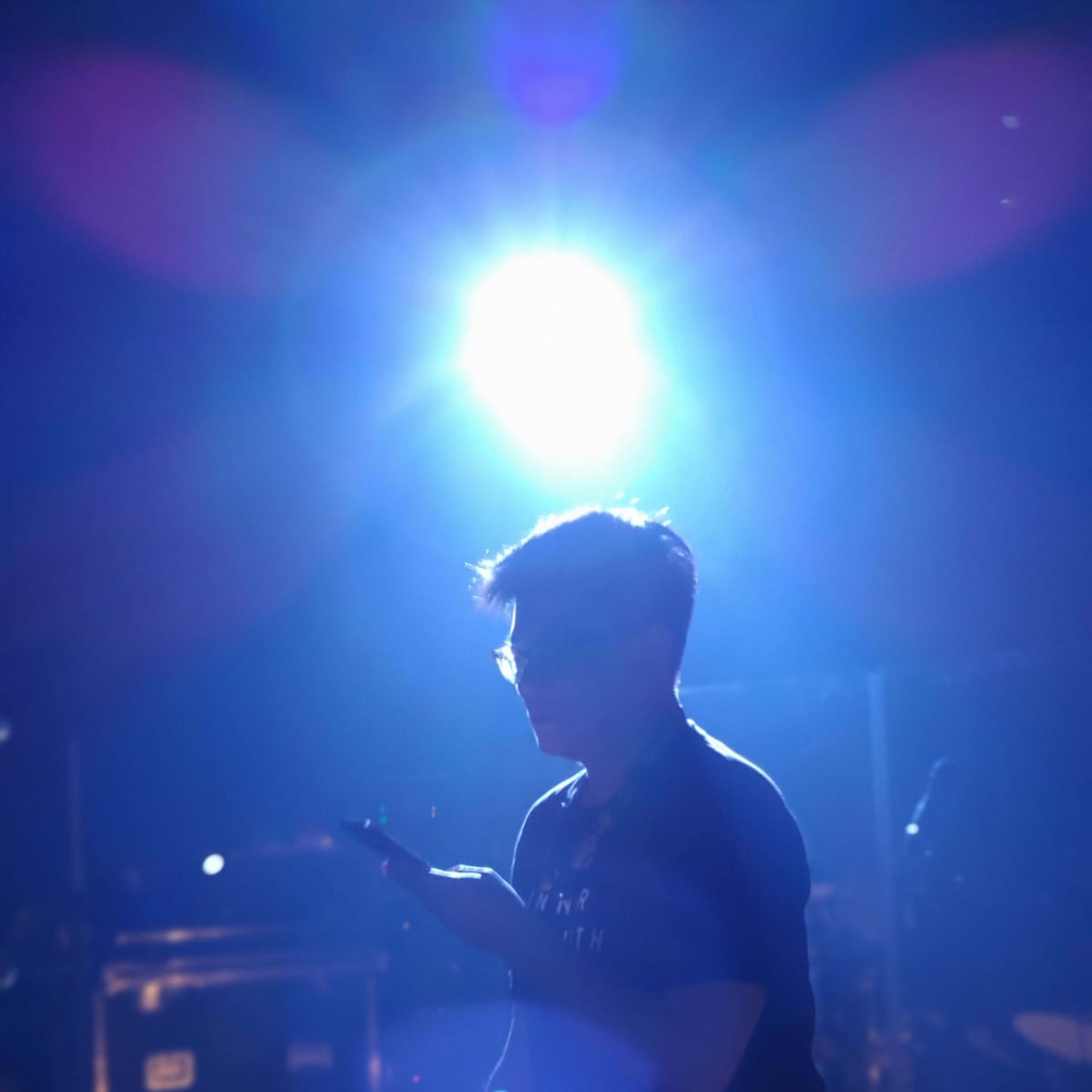A profile headshot of actor Bills Tin captured during a rehearsal for a Rooftop Productions (天台製作) performance. He is sitting on a stool under a dramatic, bright blue spotlight, looking down at a mobile device held in his hands. The atmospheric lighting creates a sharp silhouette and a lens flare effect, typical of contemporary Hong Kong experimental stage productions.